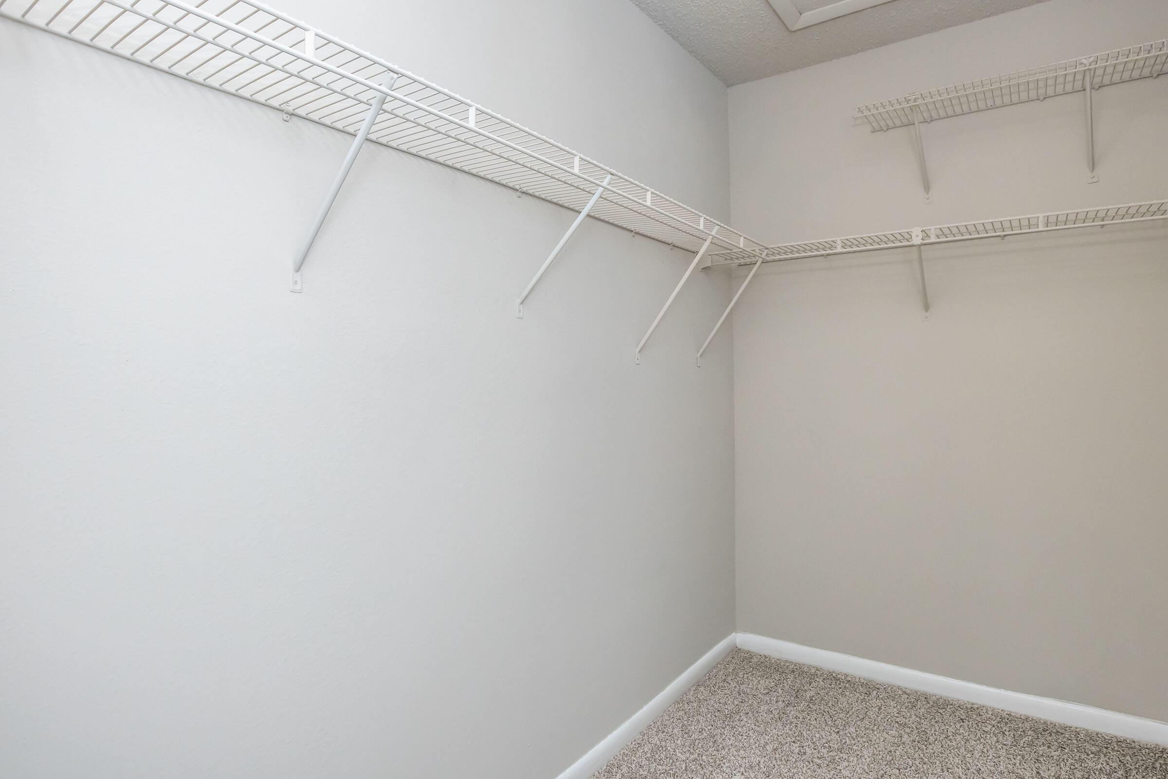 A vacant closet with light gray walls and a carpeted floor. There are white wire shelves mounted on two sides, and the space is empty, providing a clean and organized look.
