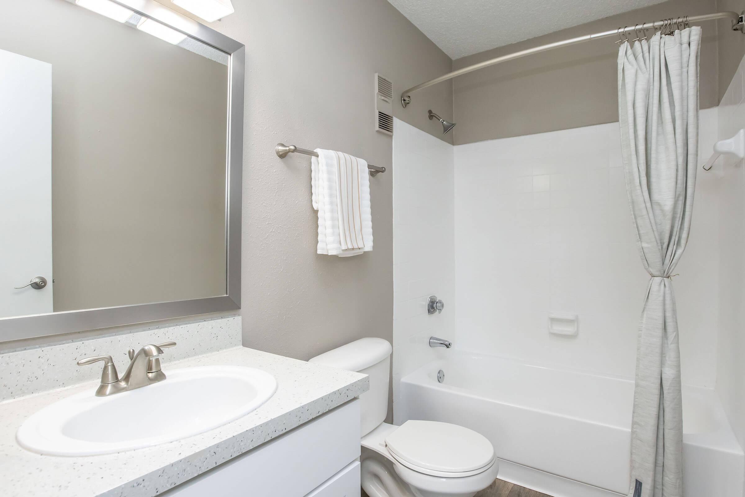 A clean, modern bathroom featuring a bathtub with a shower curtain, a white sink with a faucet, and a large mirror above the sink. Light gray walls complement the neutral decor, and there are towels neatly hung on a rail. The overall space appears bright and well-maintained.