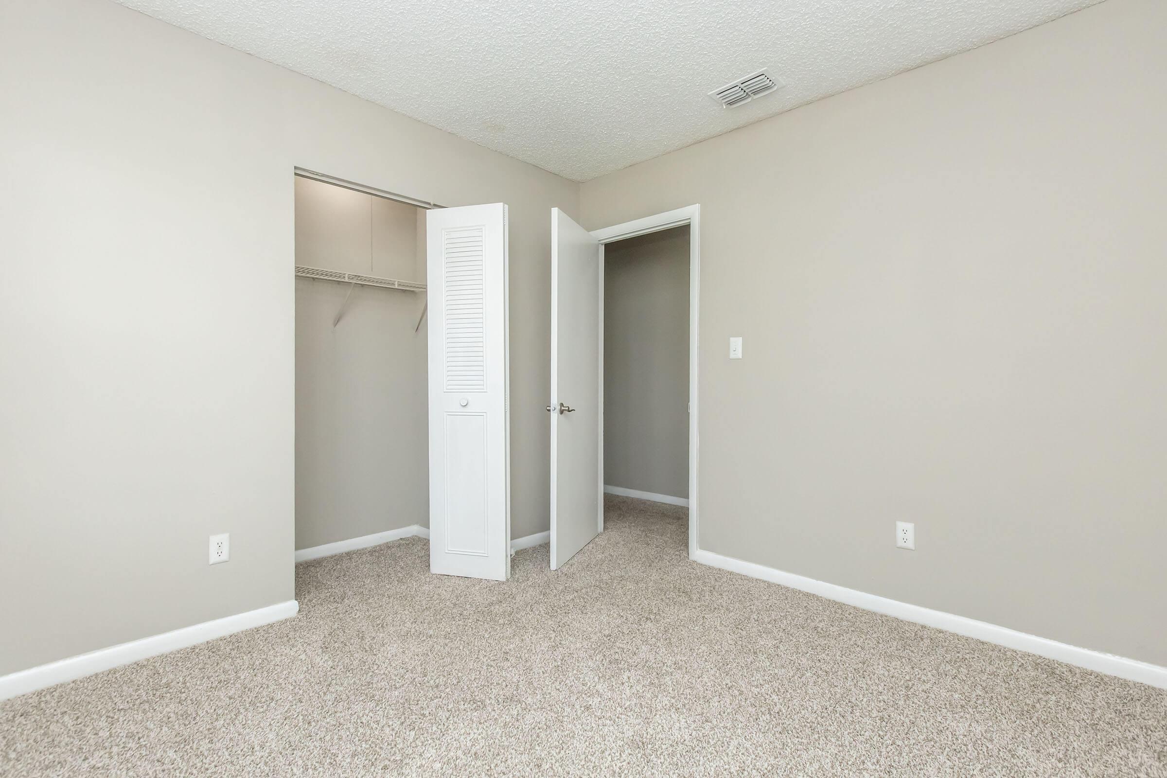 A spacious, empty room with beige walls and light carpet. There's an open door leading to a closet on the left and a door on the right, both with white trim. Natural light gently illuminates the space, emphasizing the clean, uncluttered appearance, making it suitable for various setups or decorating styles.