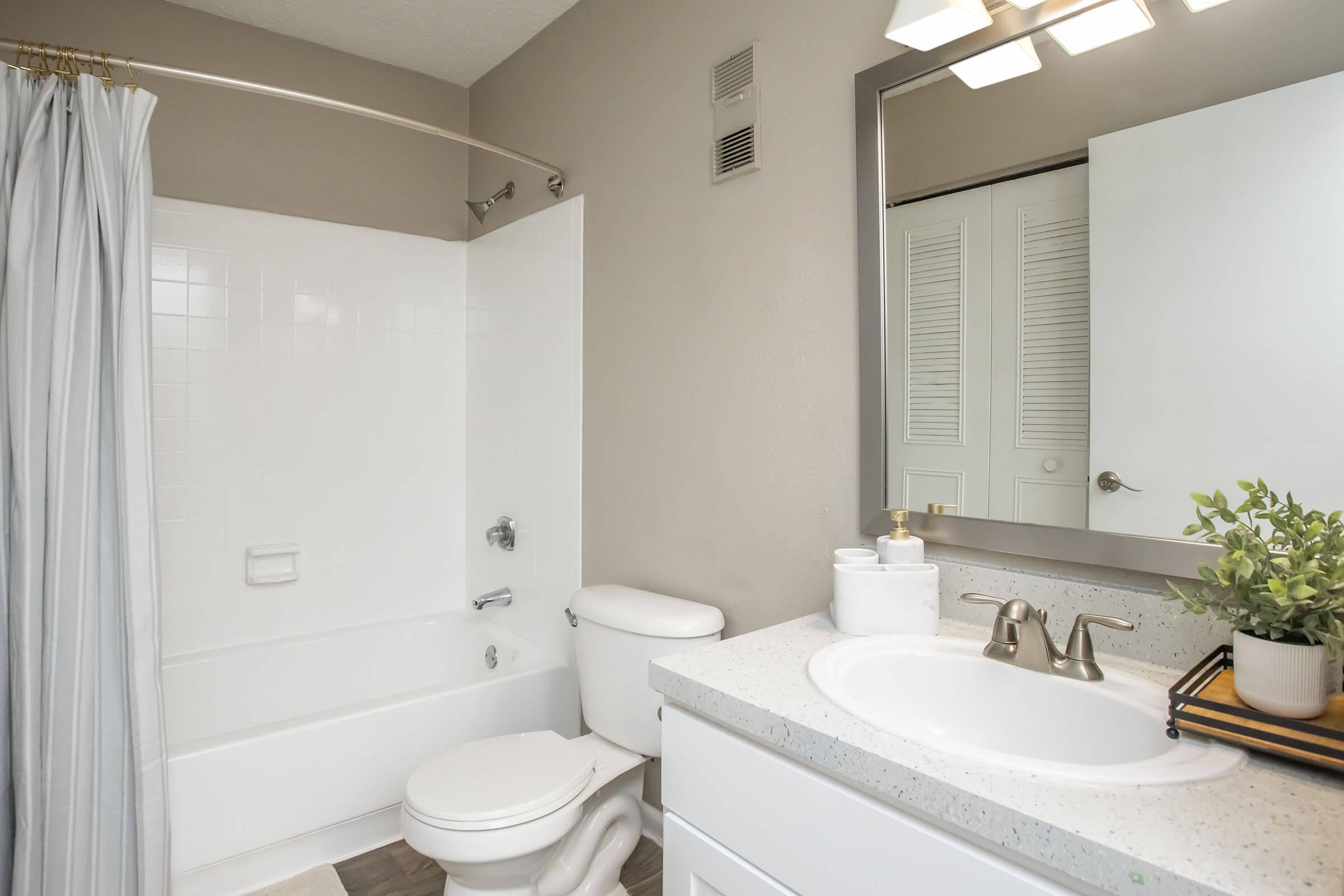 A clean, modern bathroom featuring a bathtub with a shower curtain, a toilet, and a sink with a countertop. The walls are painted in a soft neutral color, and there is a mirror above the sink. A small plant adds a touch of greenery to the countertop, enhancing the serene atmosphere.