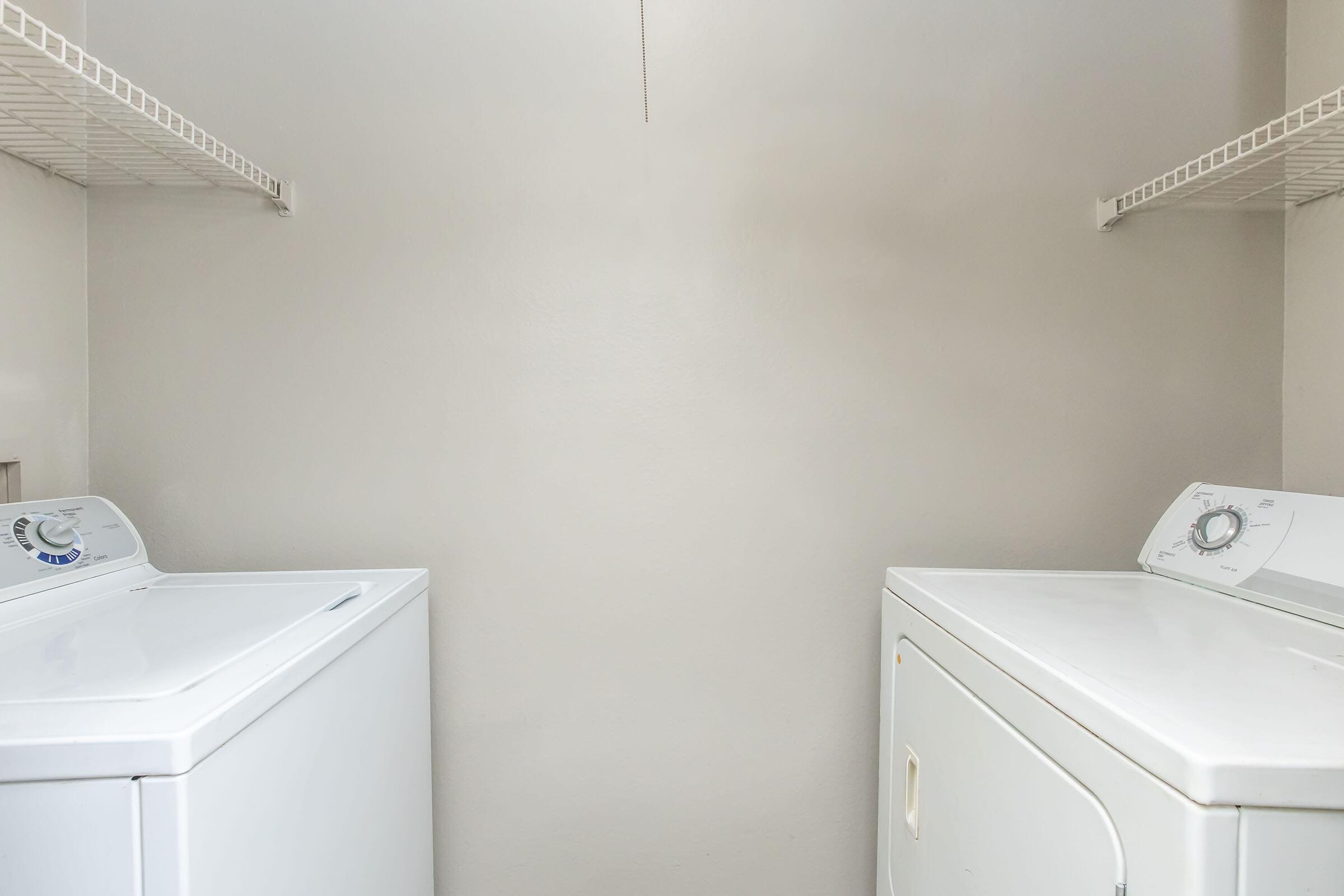 A laundry room featuring a white washing machine and a white dryer positioned side by side. The walls are painted a light color, and there are wire shelves above the appliances. The space is well-lit and organized, with no additional items visible in the room.