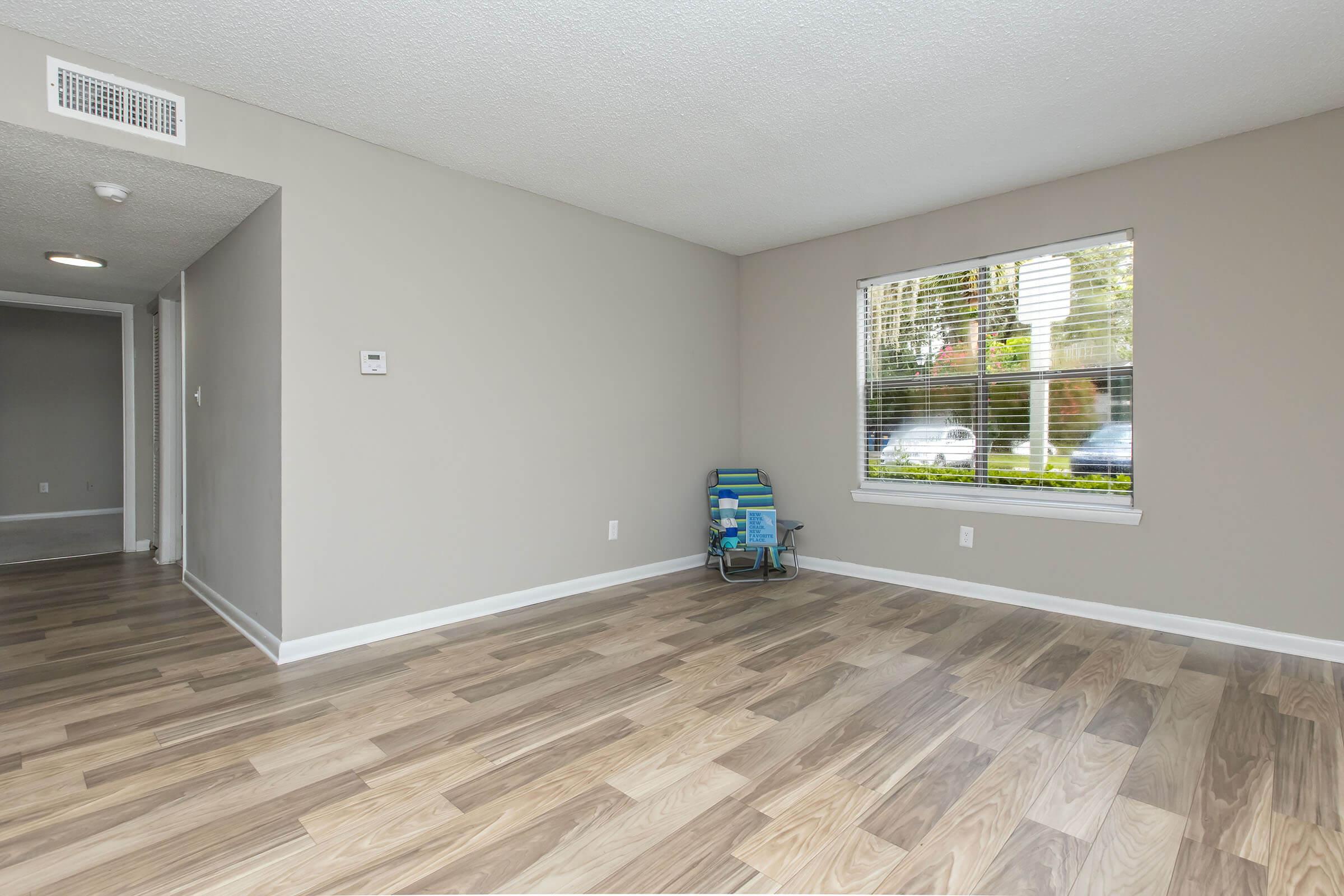 A vacant room with light-colored walls and wooden flooring. There's a window with blinds that allows natural light in, and a small blue chair in the corner. The room is organized, giving a sense of spaciousness, with a doorway visible in the background leading to another area.