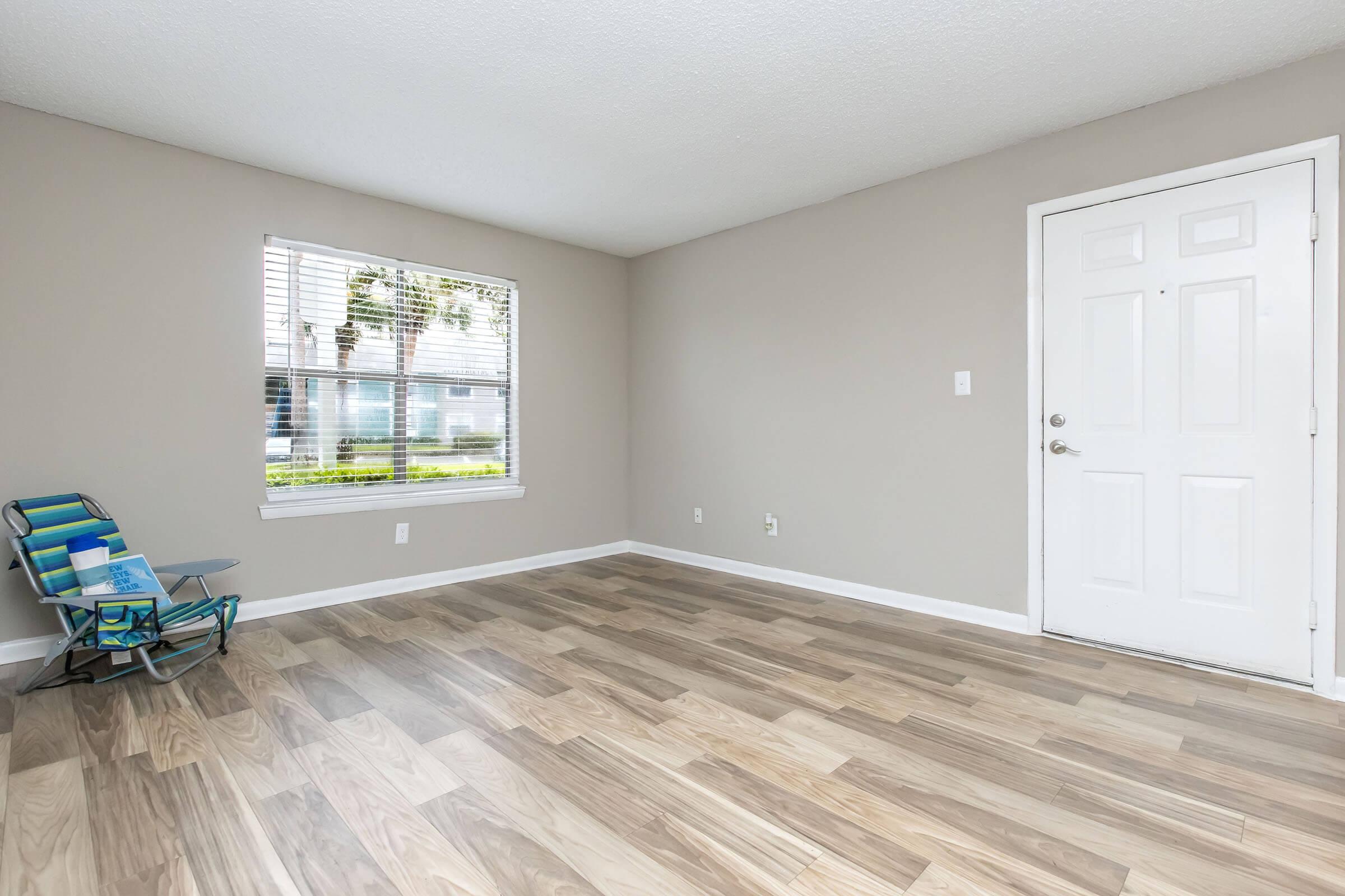 A clean, empty room with light gray walls and wood-style flooring. There is a window with white blinds allowing natural light in, and a small blue folding chair is positioned near the window. The door to the room is white and has a silver handle. The overall atmosphere is bright and minimalistic.
