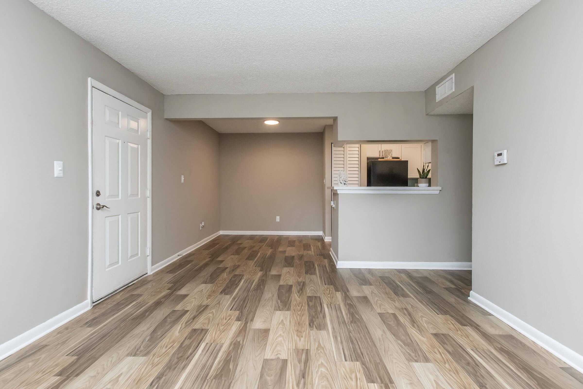 A spacious and modern living area featuring light gray walls, textured ceilings, and a wood-style floor. The room includes a front door on the left and an opening leading to a kitchenette with a small countertop. The space is well-lit and has a clean, uncluttered look, perfect for furniture arrangement.