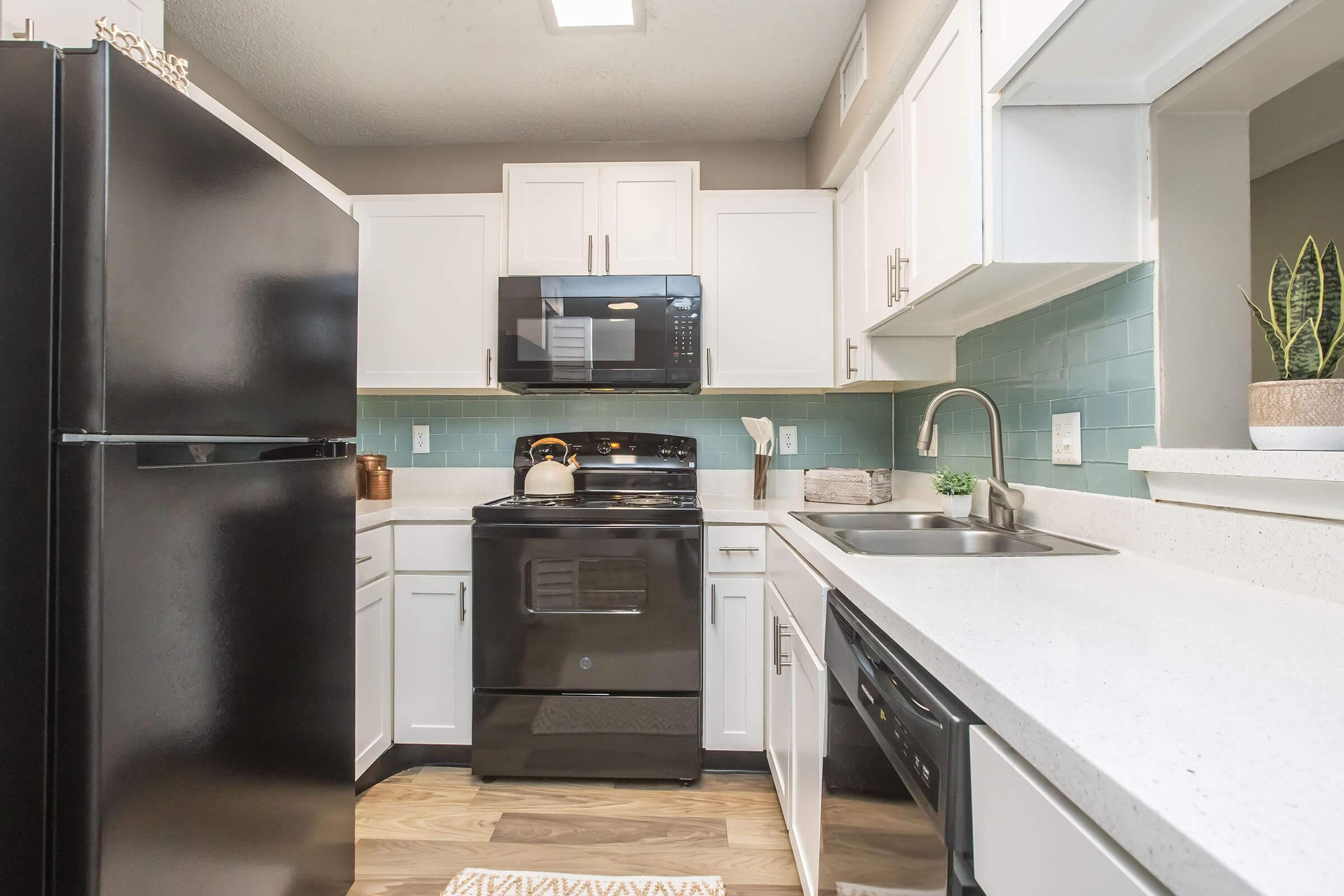 Modern kitchen featuring a black refrigerator and black stove, white cabinetry, and light blue backsplash. A microwave is installed above the stove, and a stainless steel sink is mounted on the countertop. A small plant sits on the counter, and the floor is finished in light wood tones.