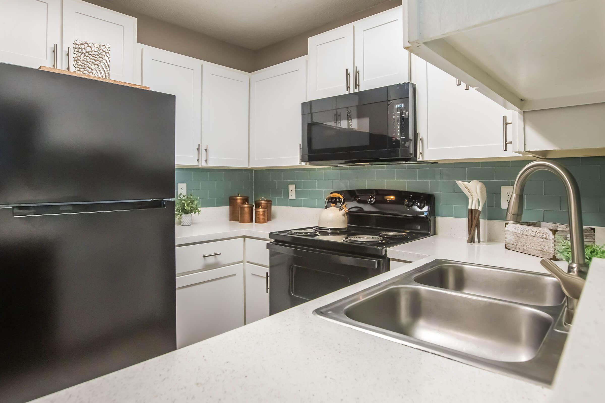 Modern kitchen featuring a black refrigerator, white cabinets, and a stainless steel sink. A black microwave is mounted above the stove, which has a kettle on it. The backsplash is adorned with teal tiles, and there are copper pots and a few decorative items on the counter. Natural light enhances the space.