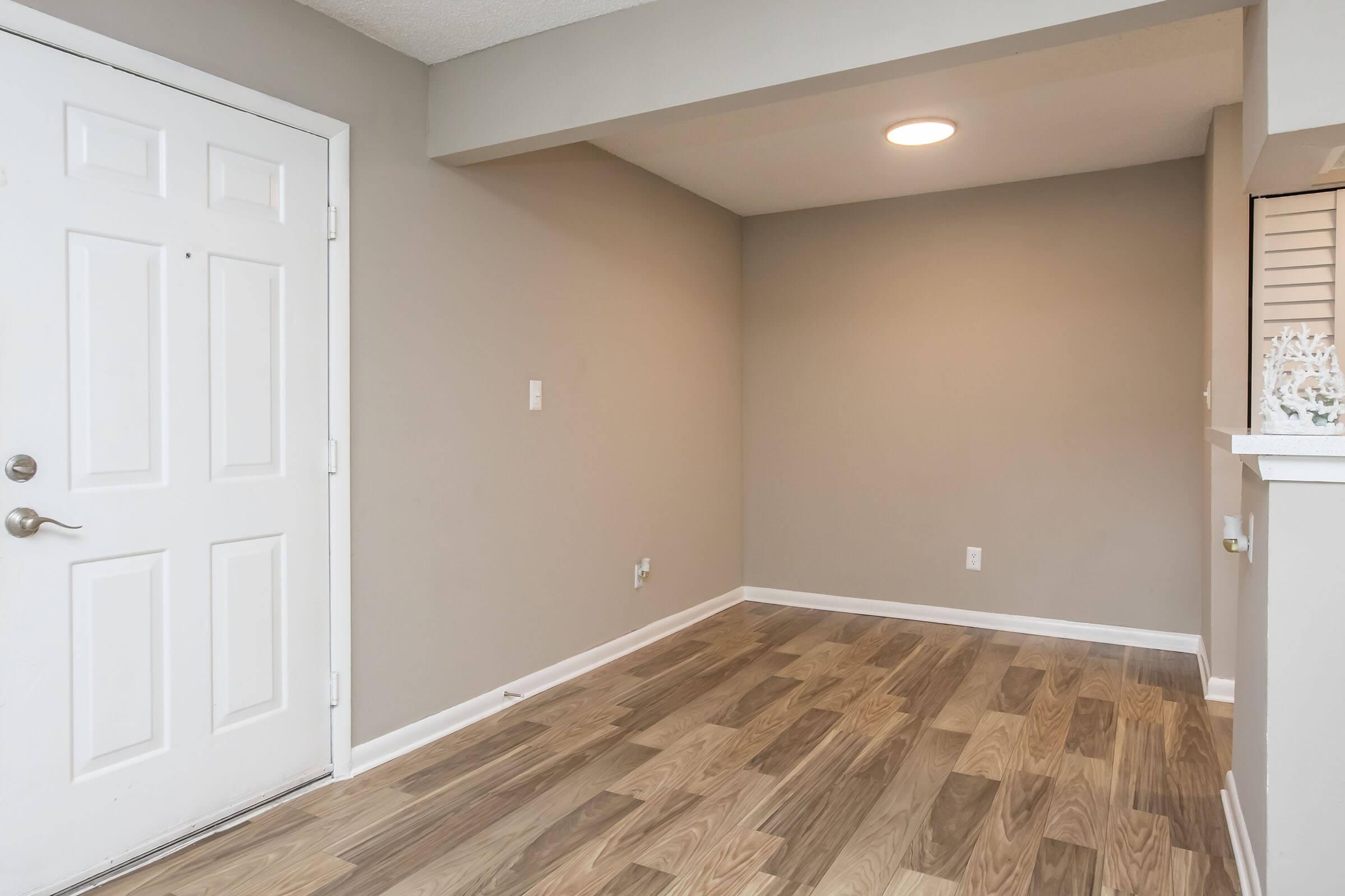 A well-lit entryway with a plain white door and light gray walls. The flooring consists of light wood laminate. The space appears empty, with a small opening to another area on the right, featuring a partial wall and some decorative elements.
