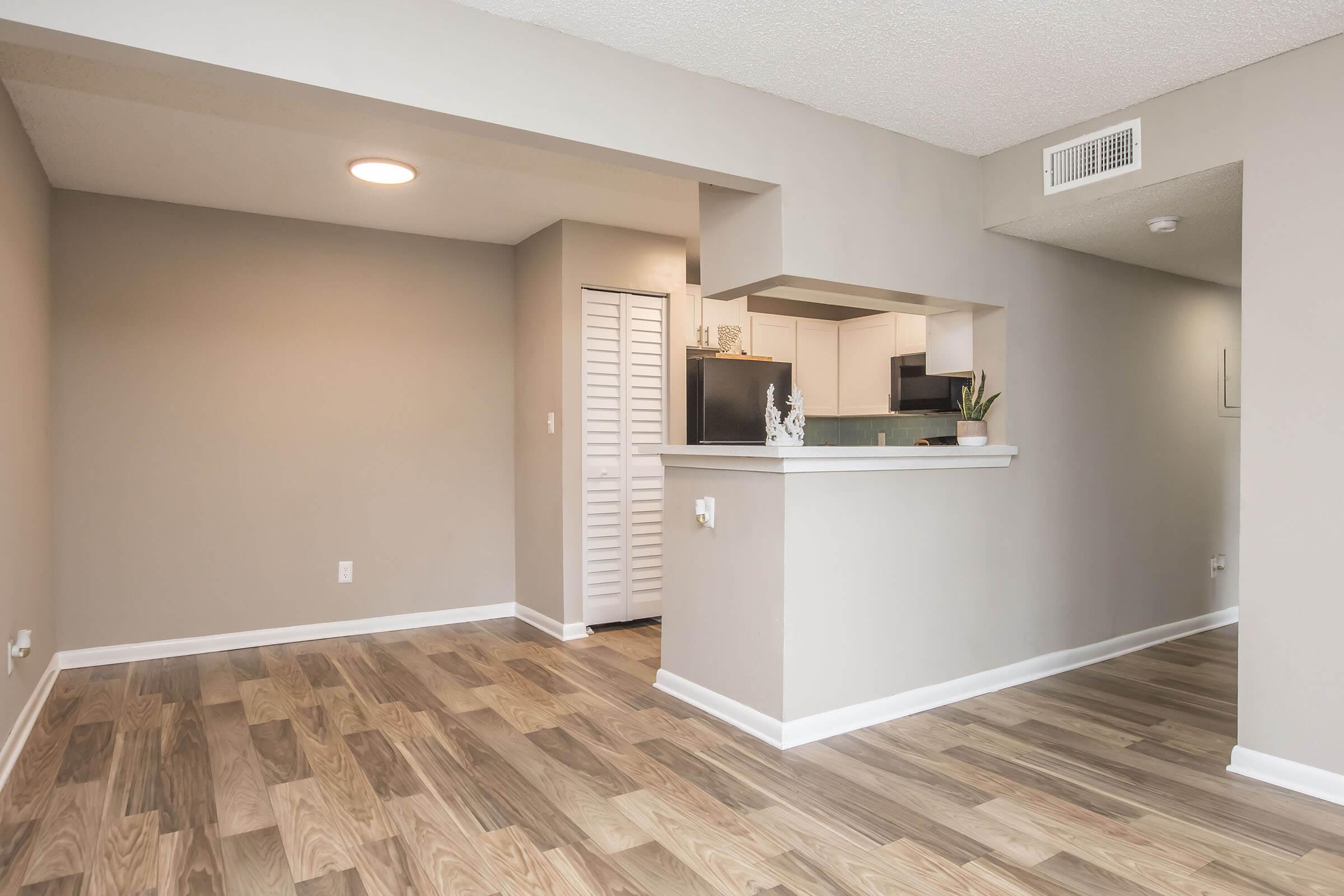 A spacious, well-lit apartment interior showcasing a neutral color palette. The area features a combination of hardwood and tile flooring. To the left, a doorway leads to a closed room, while the open kitchen is visible in the background. Natural light enhances the inviting atmosphere of the living space.