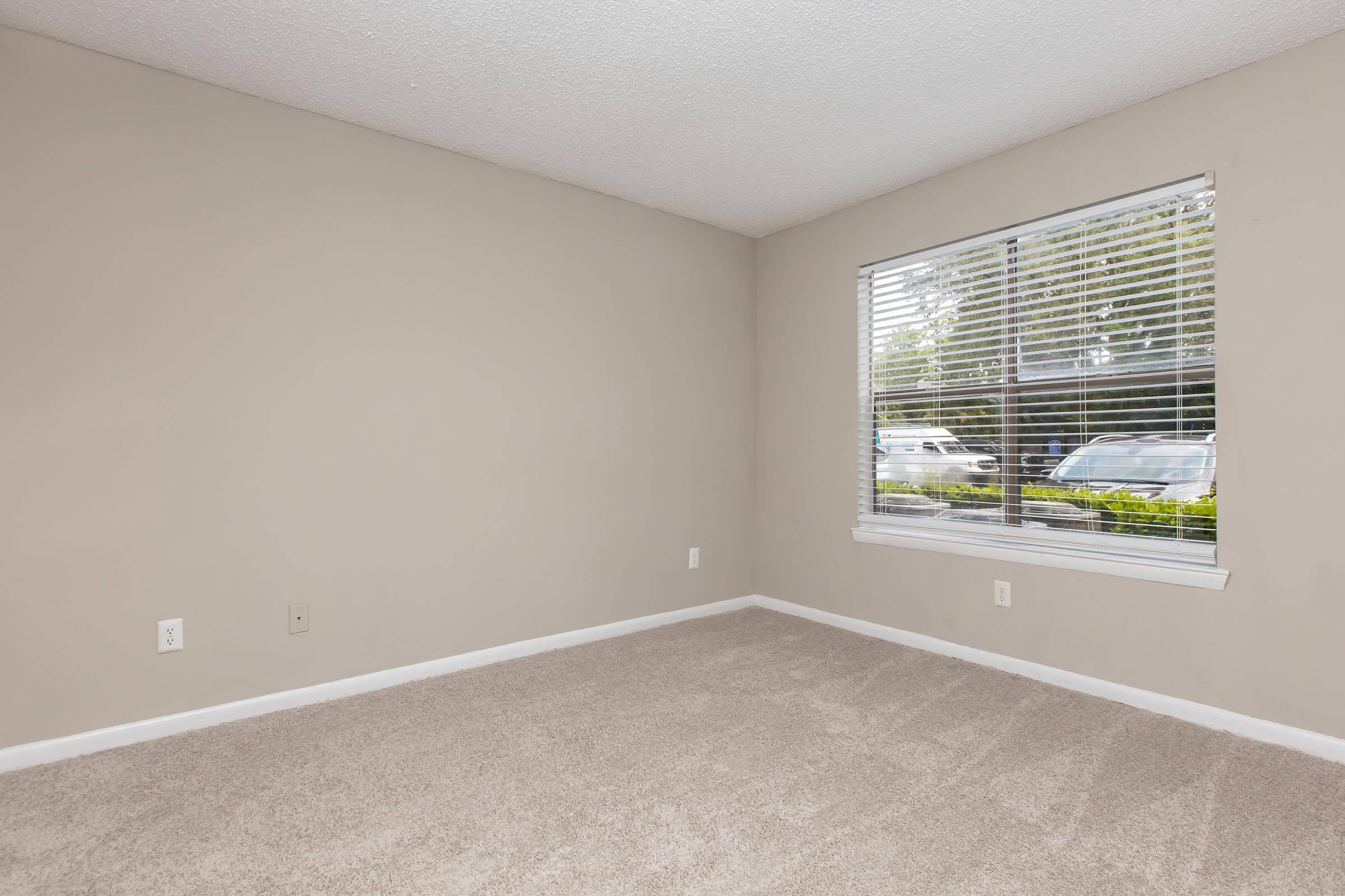 Empty room with light beige walls and carpet. A window with white blinds lets in natural light, revealing a view of green foliage and parked cars outside. The space is devoid of furniture, creating a clean and open atmosphere suitable for various uses.