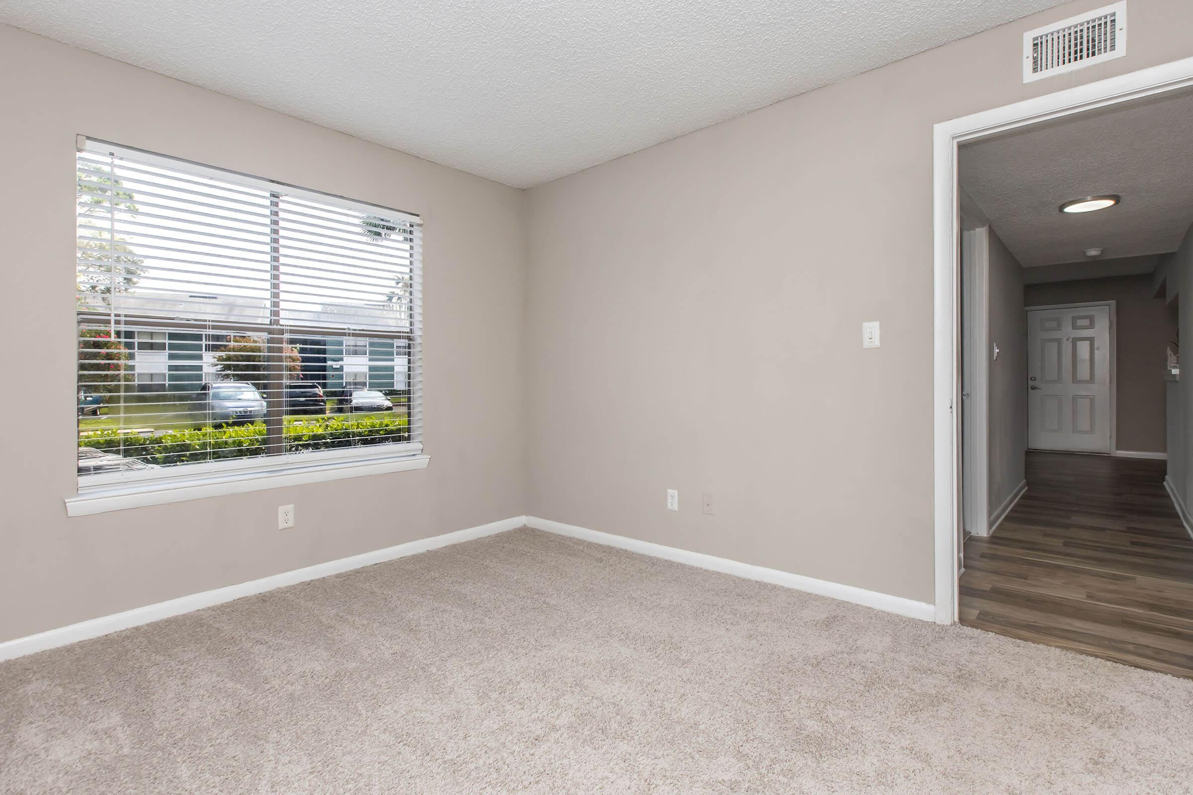 Empty room with light beige walls and gray carpet. A window with blinds allows natural light to enter, and the view outside shows greenery. A hallway is visible on the right, leading to another part of the space. The overall ambiance is bright and spacious, suitable for various uses.