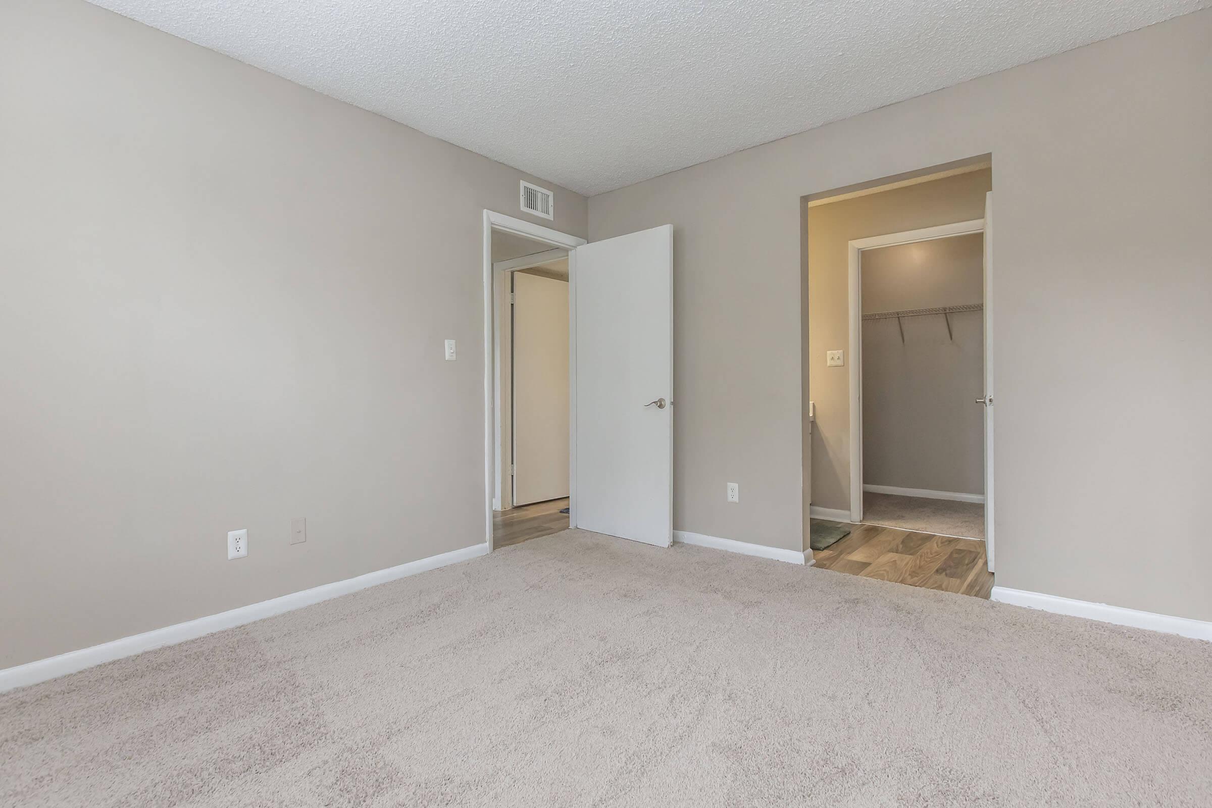 A spacious, empty bedroom with beige walls and carpet. There are two doorways—one leading to a bathroom with a coat rack visible, and another leading to an adjacent room. The room is well-lit, featuring a textured ceiling. The overall ambiance is clean and neutral, suitable for personal decoration.