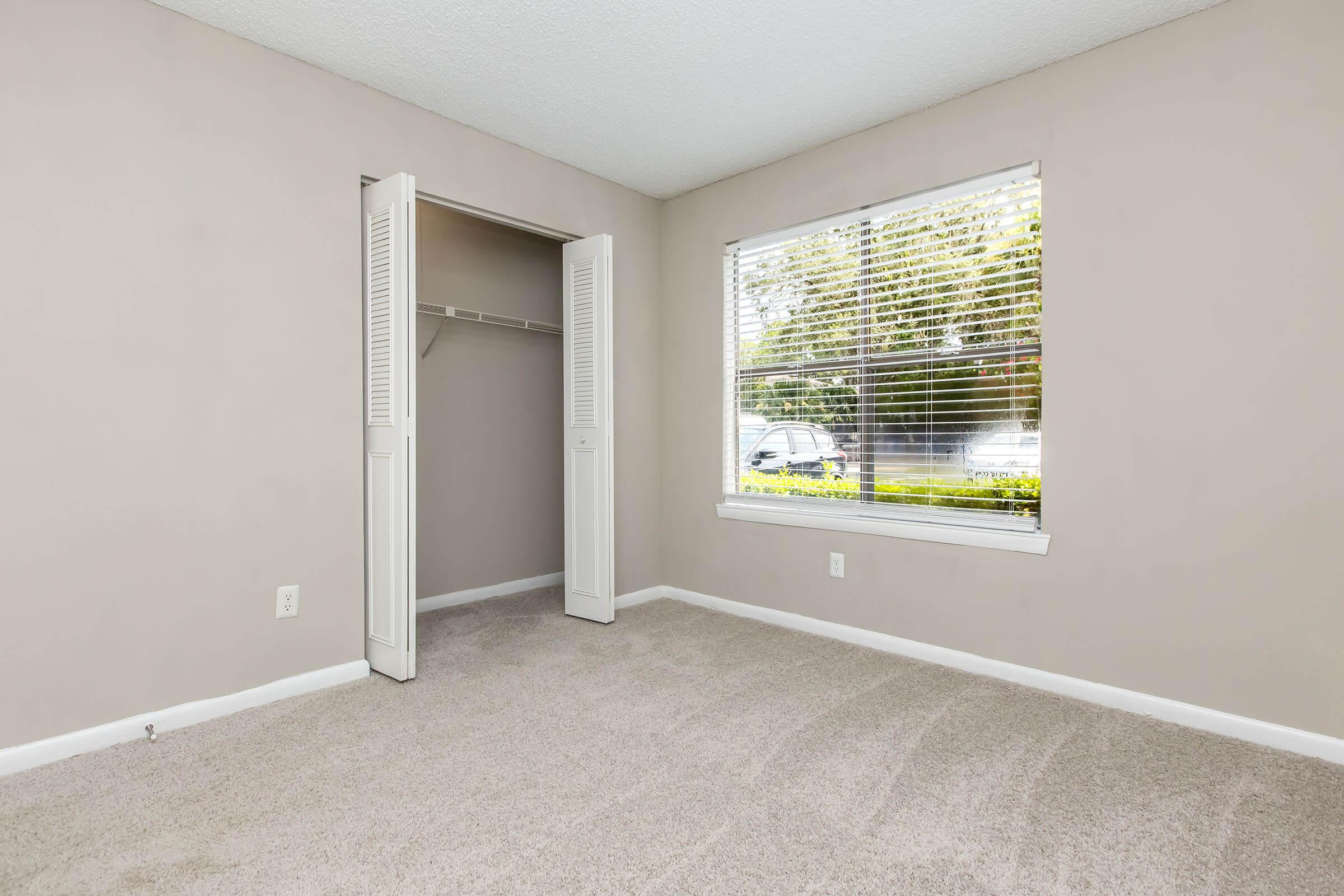 A spacious, unfurnished room with light beige walls and carpet. There's a window with blinds letting in natural light, and a small closet with sliding doors. Outside the window, greenery and a glimpse of a parked car are visible, creating a bright and airy atmosphere.