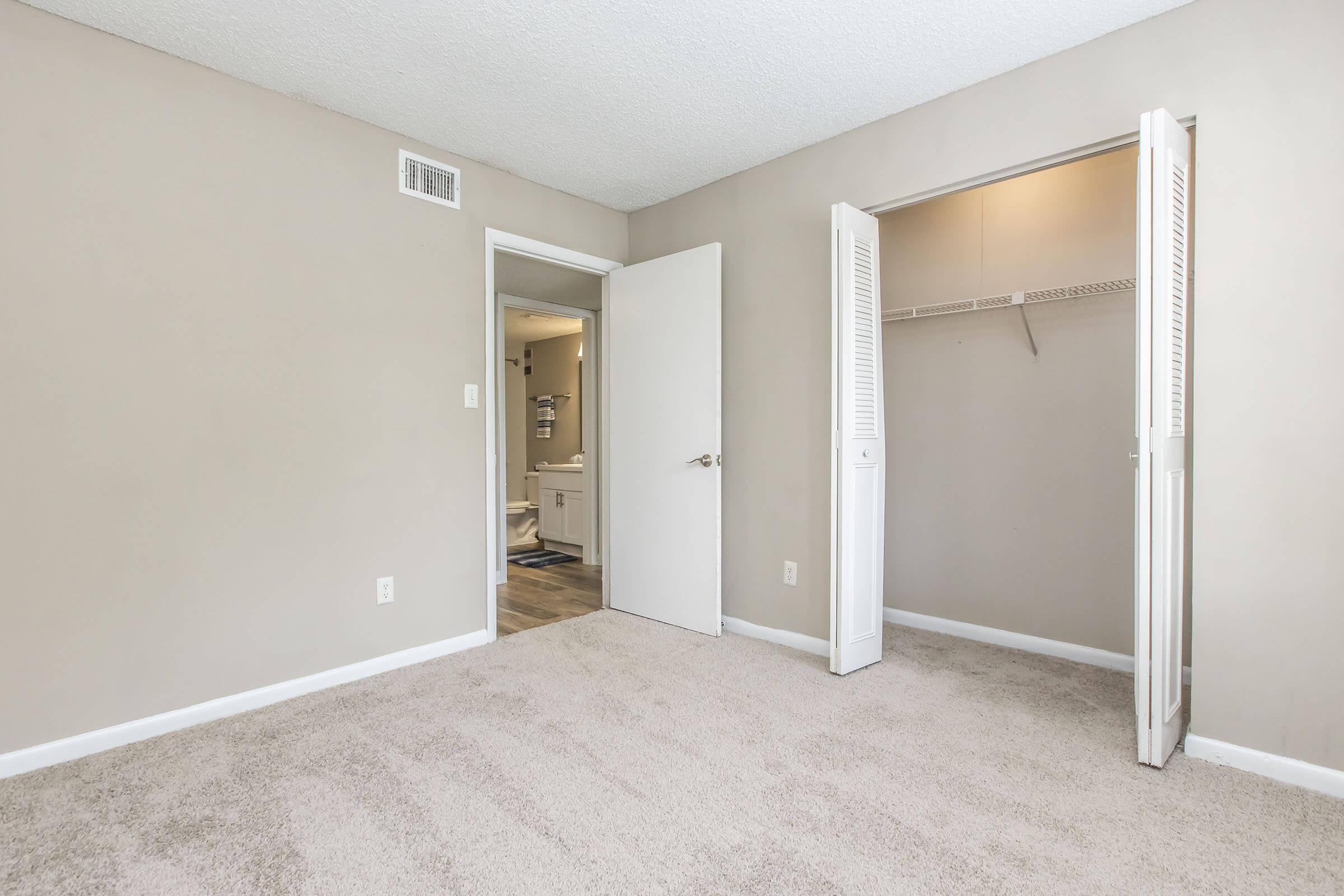 A spacious, empty bedroom with light beige walls and plush carpet. A door on the left leads to a bathroom, and there is an open closet with sliding doors on the right. Natural light brightens the room, creating a clean and airy atmosphere.