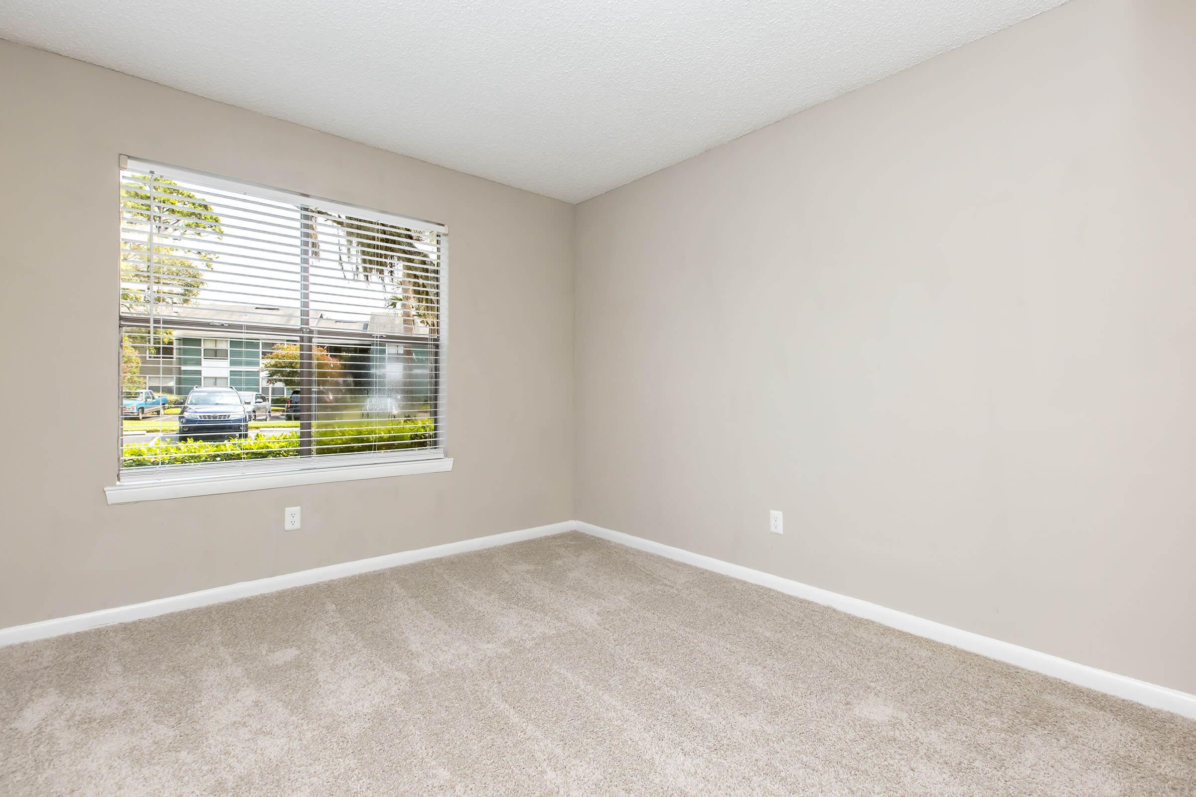 A clean, empty room with light beige walls and carpet. A window with white blinds allows natural light to enter, offering a view of greenery outside. The space appears fresh and ready for furniture placement, highlighting a minimalist aesthetic.