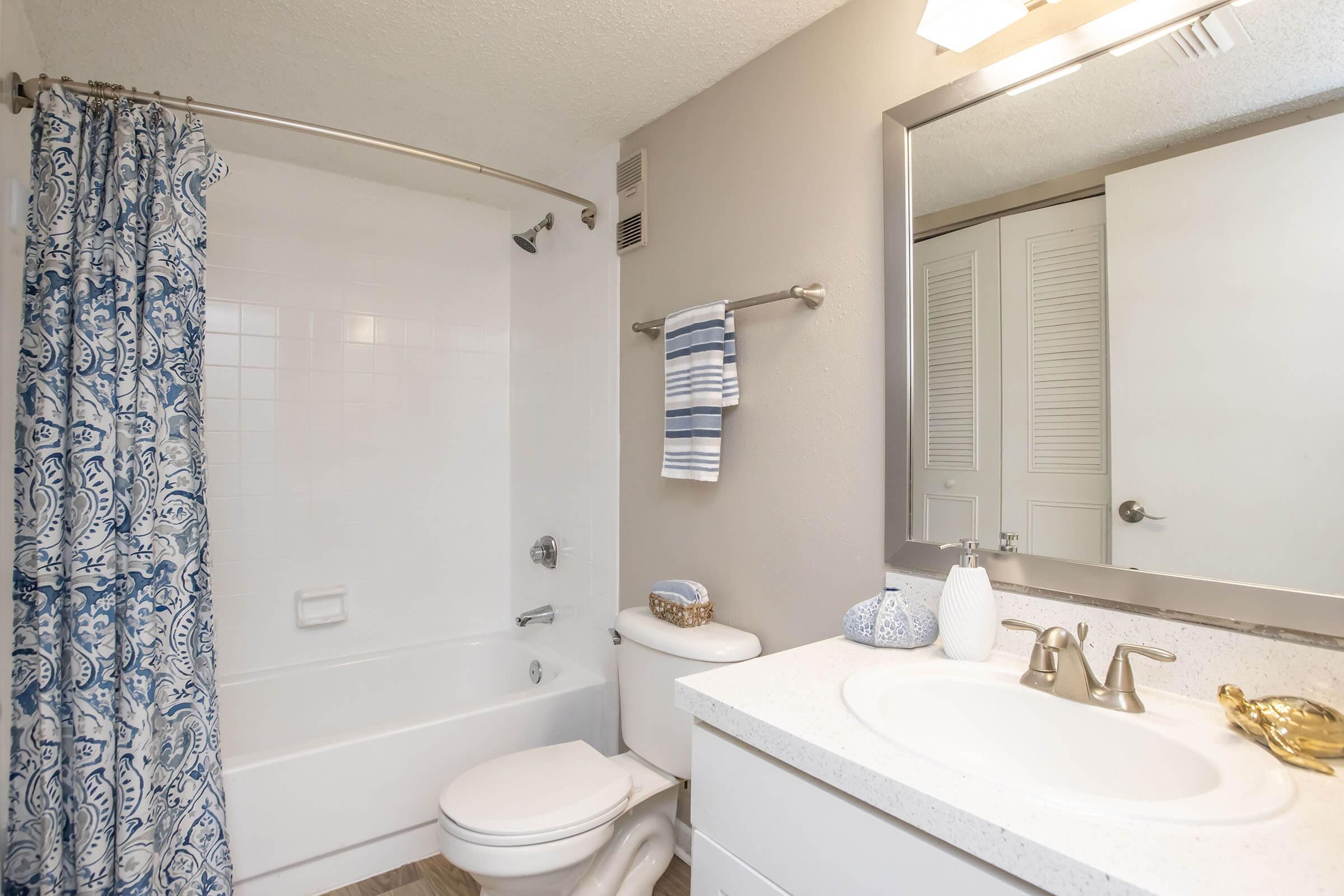A clean and modern bathroom featuring a white bathtub and shower with a blue patterned curtain, a toilet, and a vanity with a sink. The walls are painted gray, and a large mirror is above the sink. There are decorative items on the vanity and towels hanging on a rack. The space is well-lit with a ceiling light.