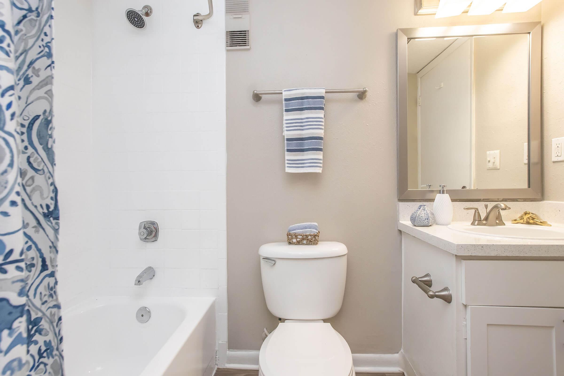 A clean and modern bathroom featuring a white bathtub with a blue and white patterned shower curtain, a wall-mounted towel rack, a toilet, and a vanity with a sink and mirror. Decorative items are placed on the countertop, and the walls are painted light gray.