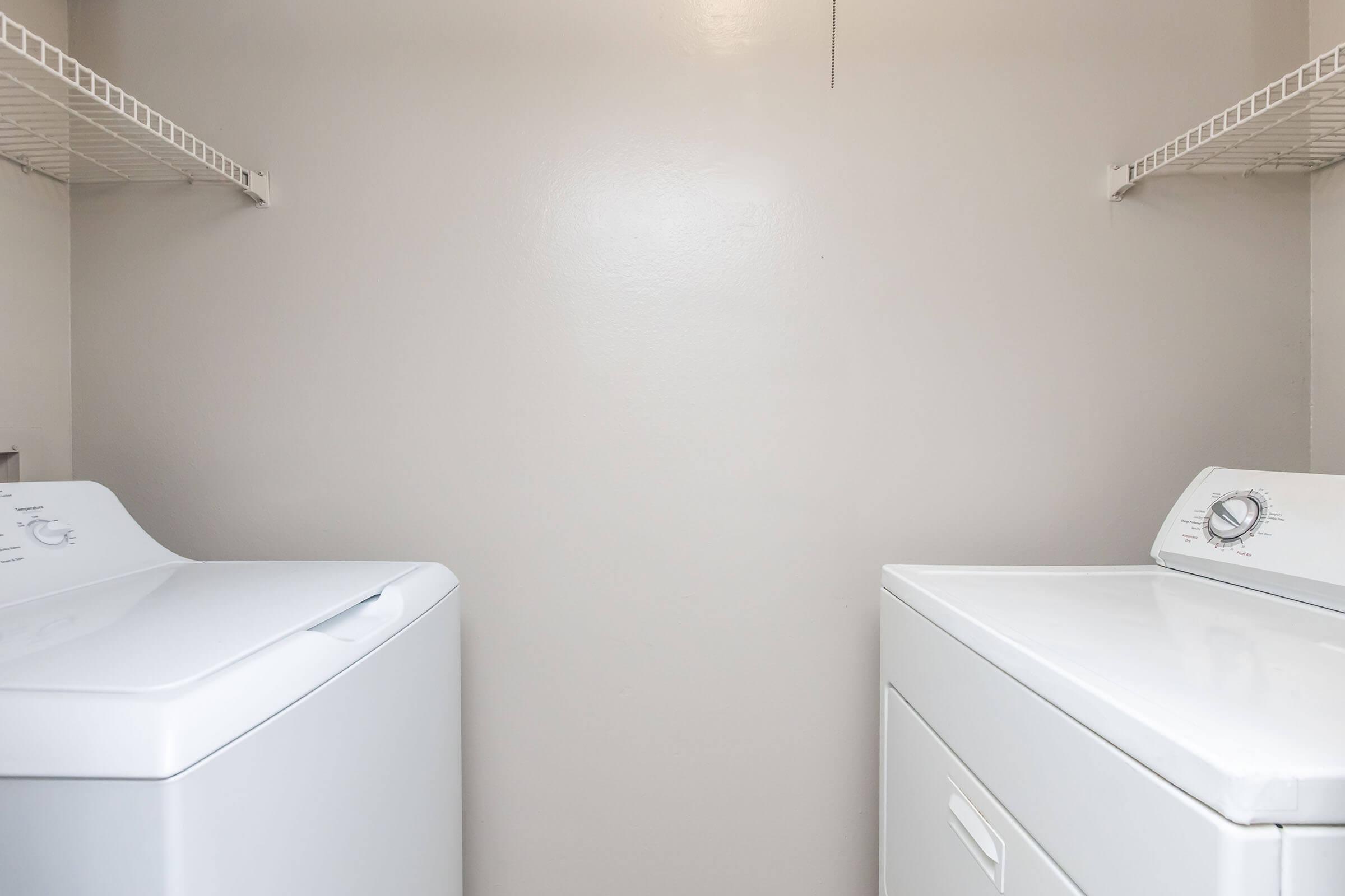 A laundry room featuring a washing machine on the left and a dryer on the right, both in white, set against a plain light-colored wall with shelving above for storage. The space appears clean and organized.