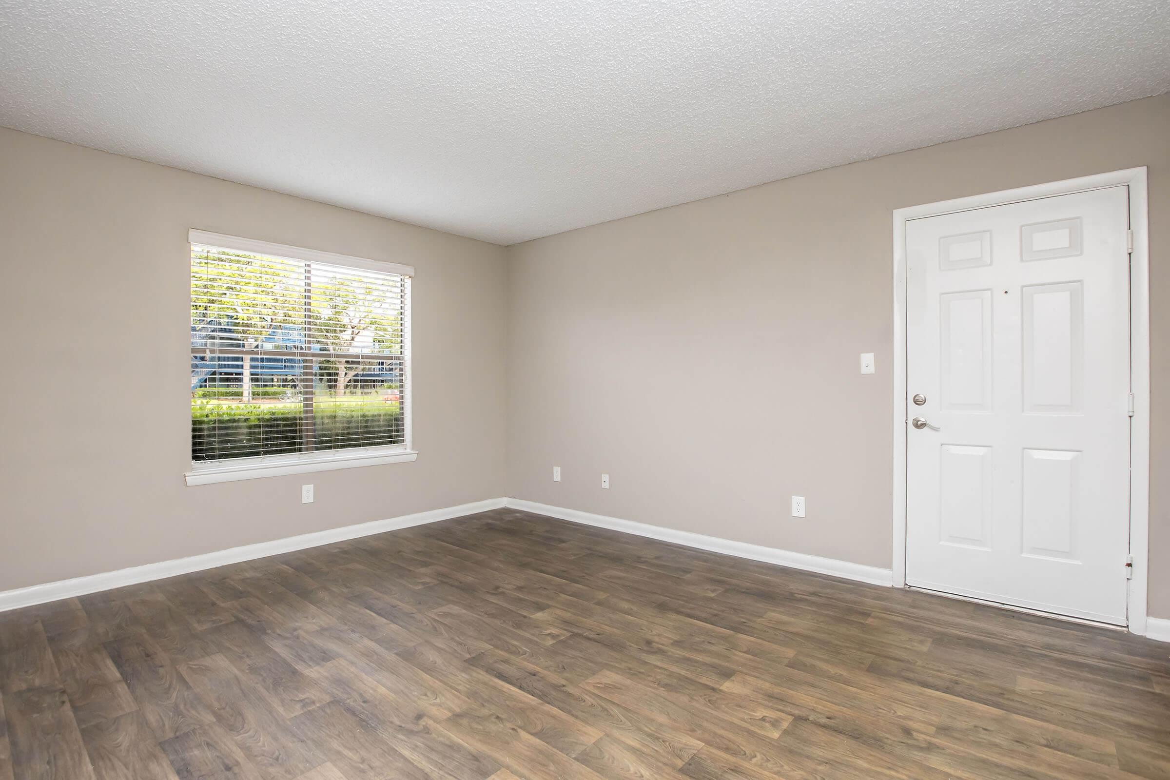 A spacious, empty room with light-colored walls and a window allowing natural light. The floor is covered in wood-like laminate. A plain white door is visible on the right, leading outside. The overall atmosphere is clean and ready for furnishings.