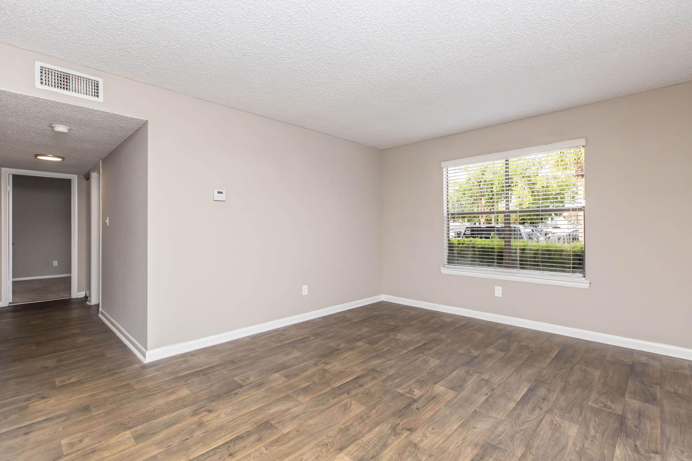 A bright, empty living space with light-colored walls and hardwood floors. A window with blinds lets in natural light, and a doorway leads to another room. The walls are smooth, and there are no furnishings, creating a clean and spacious atmosphere.