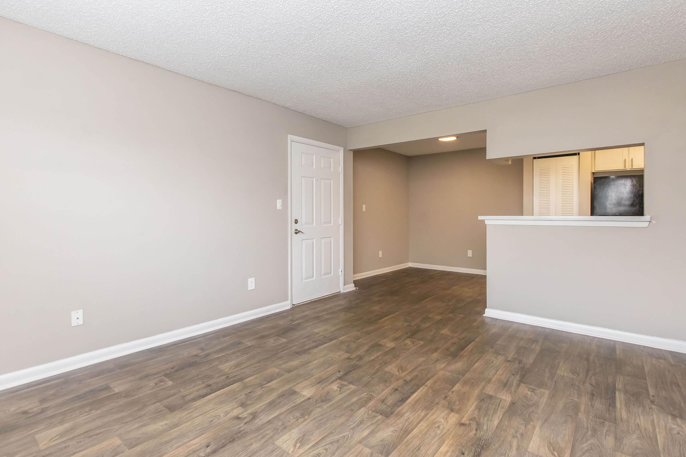 Empty living space featuring light gray walls and a wood-style floor. A white door leads outside, with an open layout showing a pass-through to a kitchen area. The room is well-lit, with a small bar counter visible, creating a spacious and inviting atmosphere.