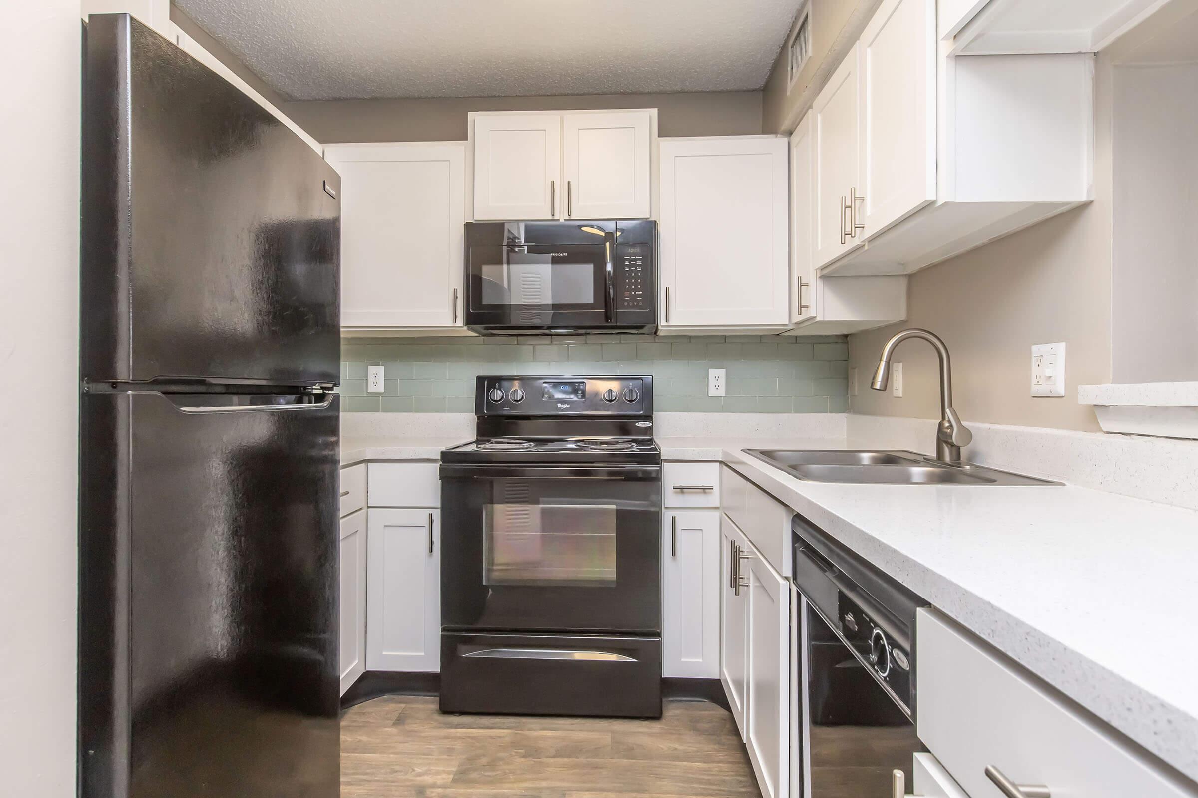 A modern kitchen featuring black appliances including a refrigerator, oven, and microwave. There are white cabinets and a light-colored countertop. A sink is positioned beside the stovetop, with green backsplash tiles adding a pop of color. The floor has a wood-like finish, giving the space a warm appearance.