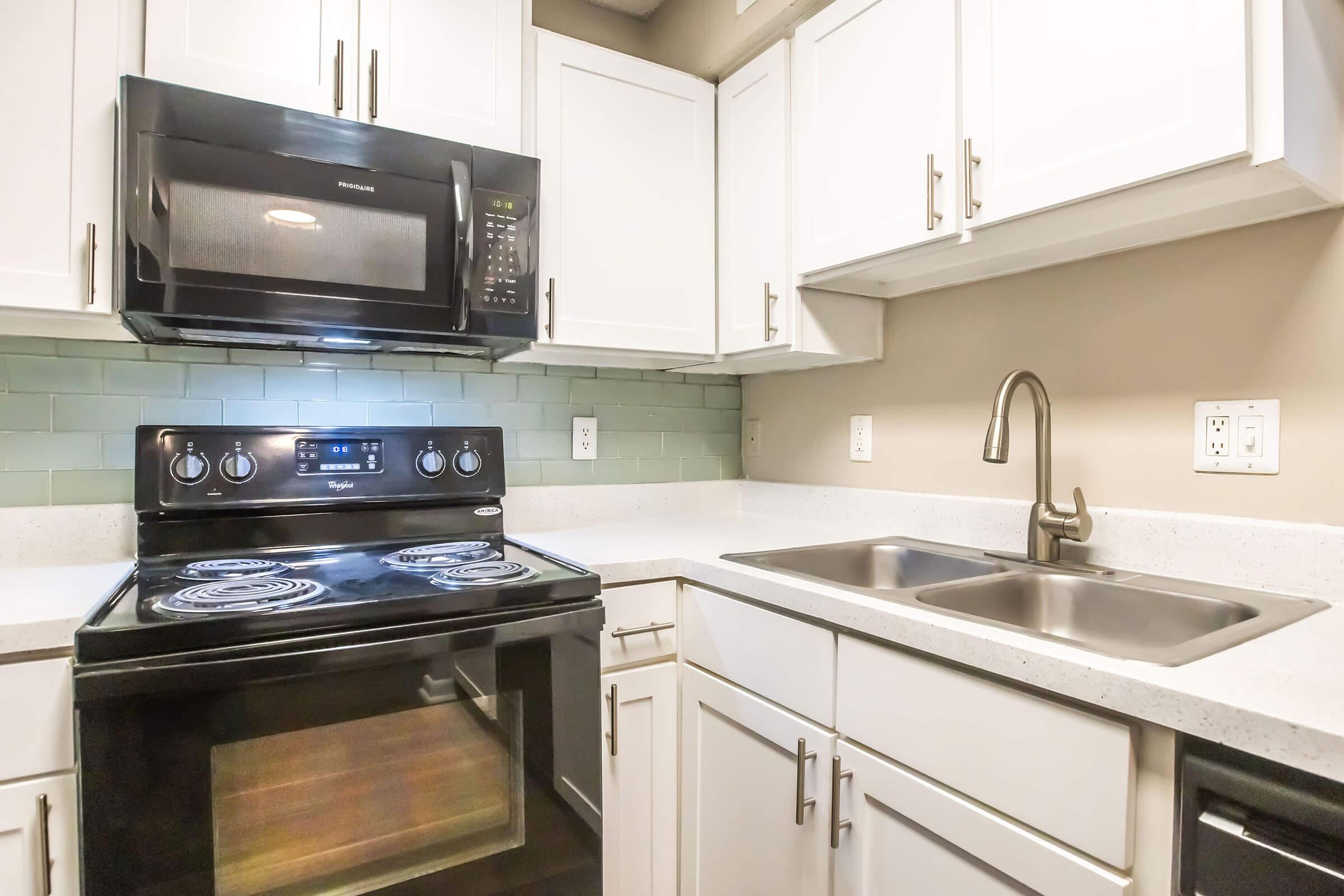 A modern kitchen featuring white cabinets, a black stovetop and oven, a black microwave, and a stainless steel sink with a brushed nickel faucet. The countertop is light-colored, and there's a green tiled backsplash behind the stovetop. The room is well-lit and organized.