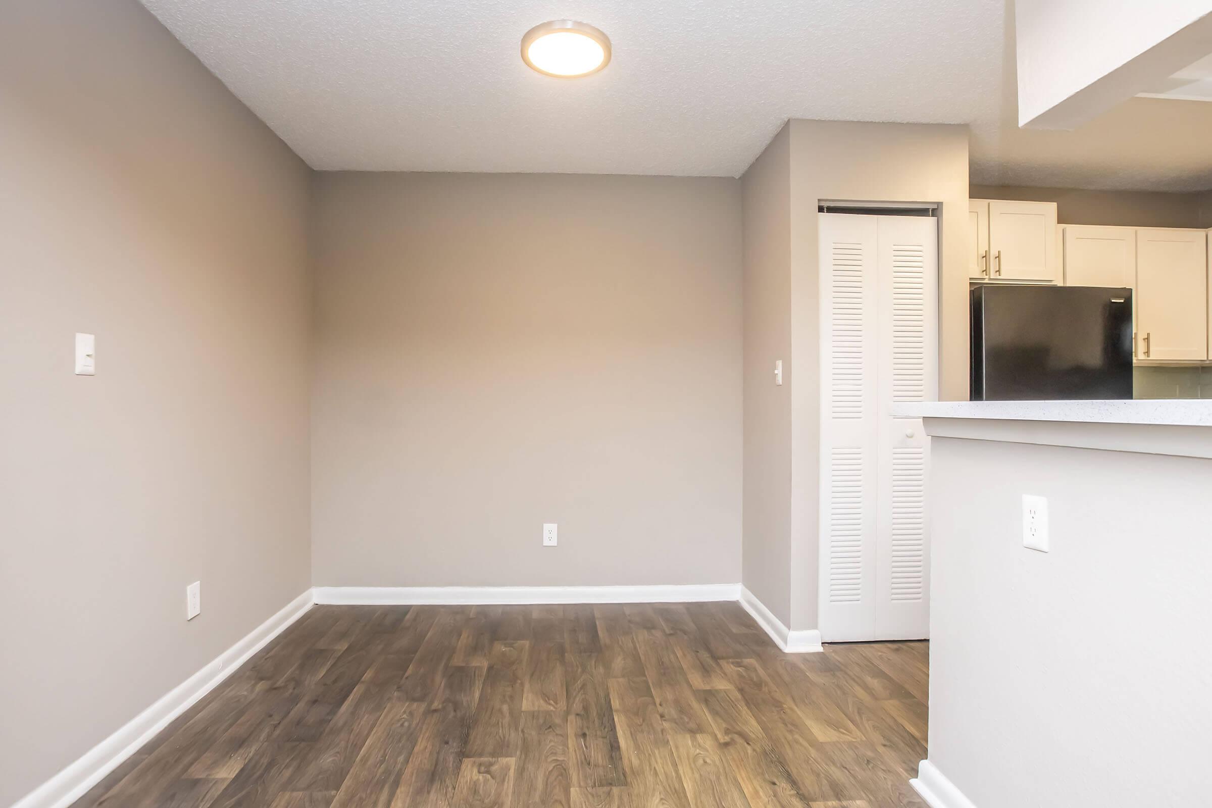 A well-lit, empty room featuring light gray walls and a wood-style floor. The room has a small wall-mounted light fixture and an open doorway leading to a closet. In the background, there is a partial view of a kitchen area with white cabinets and a black refrigerator.