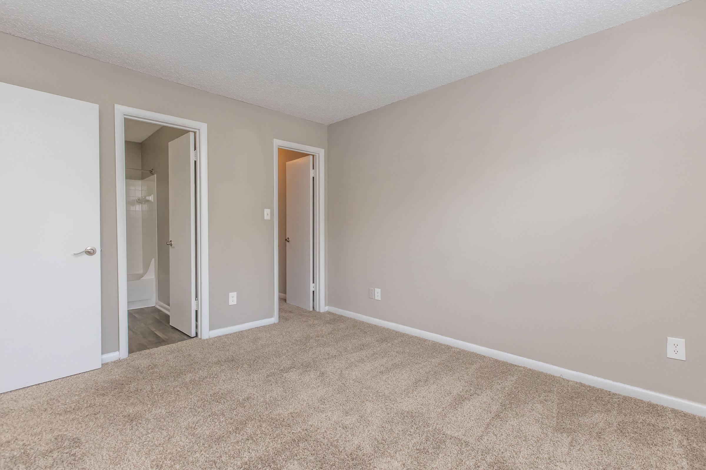 A spacious room with light beige walls and a textured ceiling. Two doorways lead to adjacent areas, with a glimpse of a bathroom visible through one of the doors. The floor is covered in soft, neutral-toned carpeting, creating a cozy atmosphere. Natural light softly brightens the space.