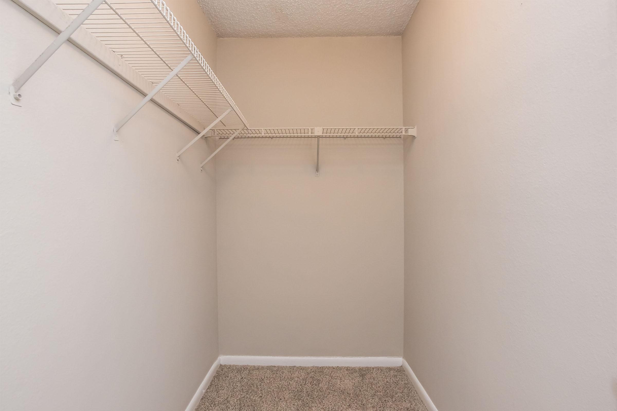 An empty closet with pale beige walls, a carpeted floor, and a metal shelving unit mounted on one side. The space is devoid of any items or furniture, emphasizing its emptiness and potential for storage.