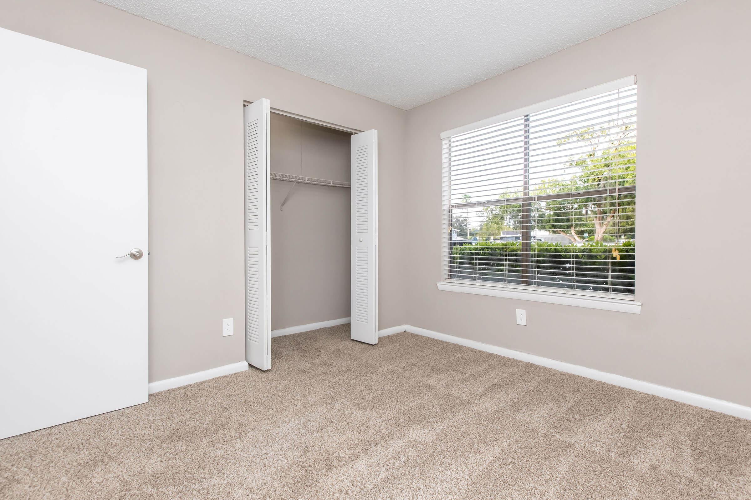 A well-lit empty room with beige walls and carpet. The room features a window with blinds on the left side, providing a view of greenery outside. On the right, there is a white door leading to a closet with sliding doors. The space is tidy and ready for new furniture or decoration.