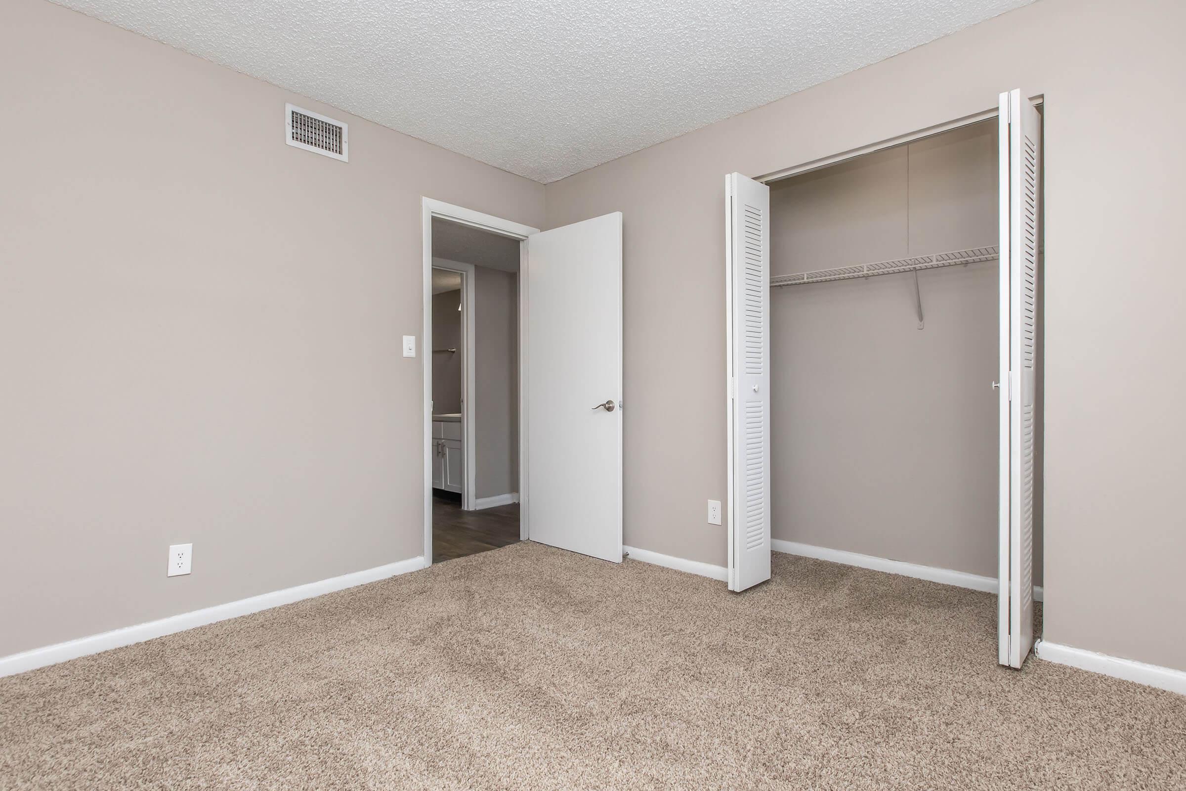 Empty room with beige walls and light carpet. A closet with white doors is open on the right, and there is a doorway leading to another room in the back. The space appears clean and well-lit, suitable for decorating or furnishing.