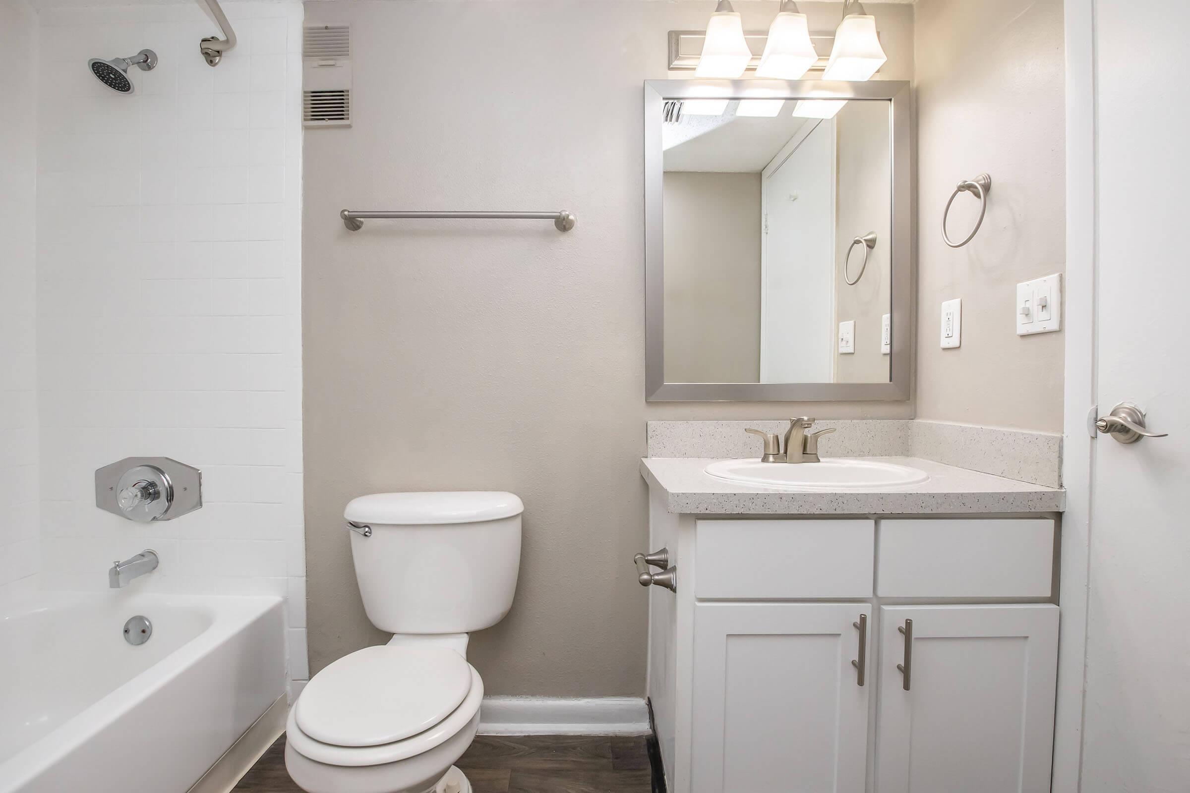 A clean and modern bathroom featuring a white bathtub with a shower, a toilet, and a vanity with a sink. The walls are painted in a light gray color, and there is a large mirror above the sink with three light fixtures. The flooring is dark, contrasting with the lighter tones of the bathroom fixtures.