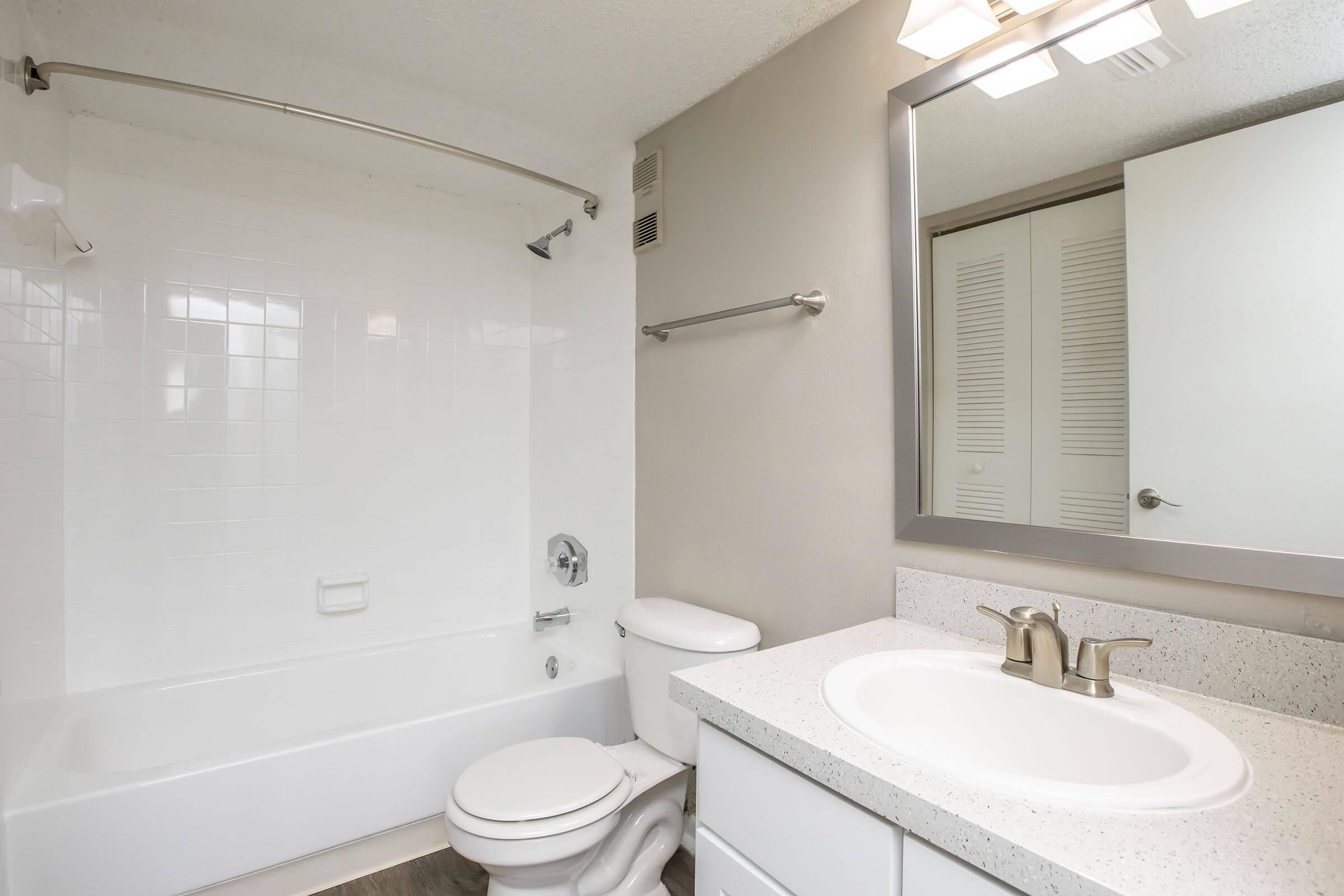 A clean and modern bathroom featuring a white bathtub, toilet, and sink with a marble countertop. The walls are painted light gray, and a mirror above the sink reflects the space. A shower curtain rod is installed, and there are storage cabinets in the background providing a minimalist look.