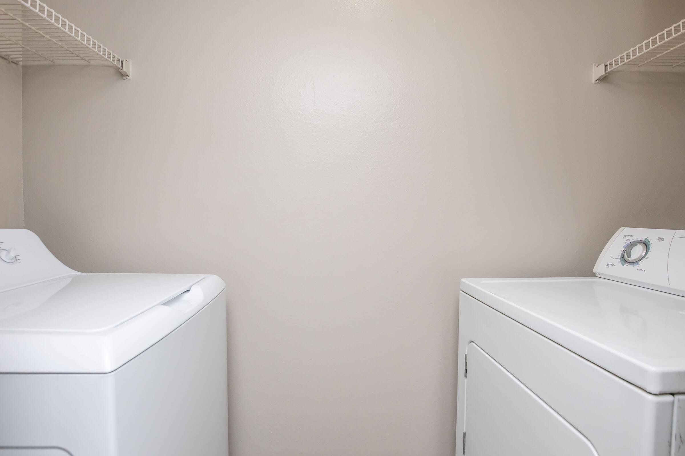 A clean and organized laundry room featuring two white appliances: a front-loading washing machine on the left and a matching dryer on the right. The walls are a soft beige color, and there are wire shelves above the machines, providing space for storage.