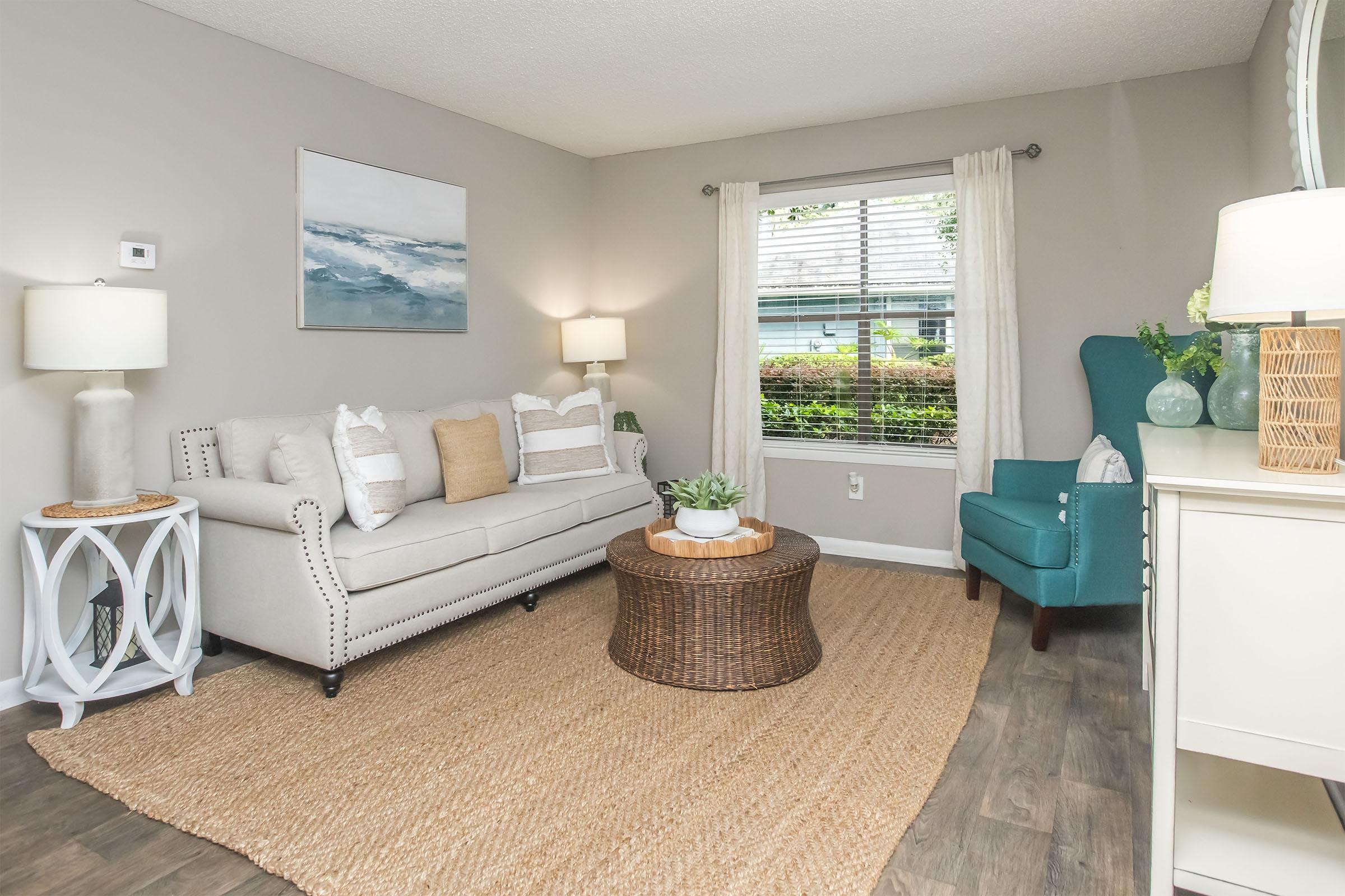A cozy living room featuring a light gray wall, a beige sofa with decorative pillows, a round wicker coffee table, and a teal armchair. Natural light streams in through a window adorned with sheer curtains, while a jute rug covers the floor. A small potted plant and lamps provide a welcoming ambiance.