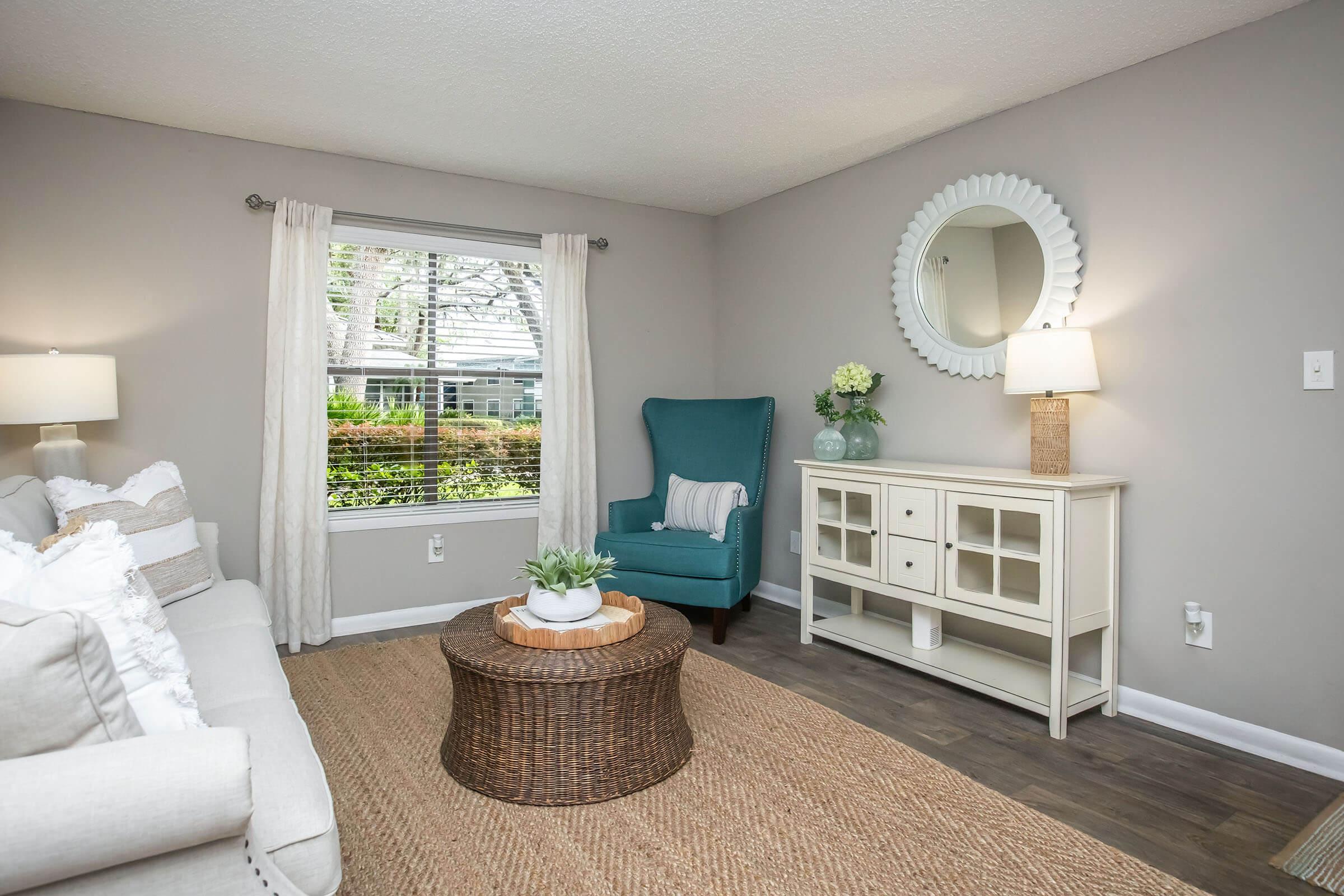 A cozy living room featuring a light gray wall, a beige sofa adorned with cushions, a round wicker coffee table, and a blue accent chair. There’s a stylish white cabinet, a soft area rug, and natural light streaming through a window, complemented by green plants and decorative items.
