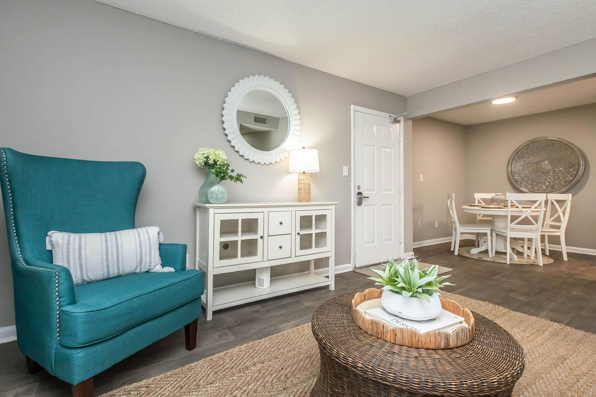 A cozy living room featuring a teal wingback chair, a round mirror above a white side table, and a modern dining area with a round table and white chairs in the background. Natural lighting enhances the neutral walls and hardwood-style flooring, creating a warm and inviting atmosphere.