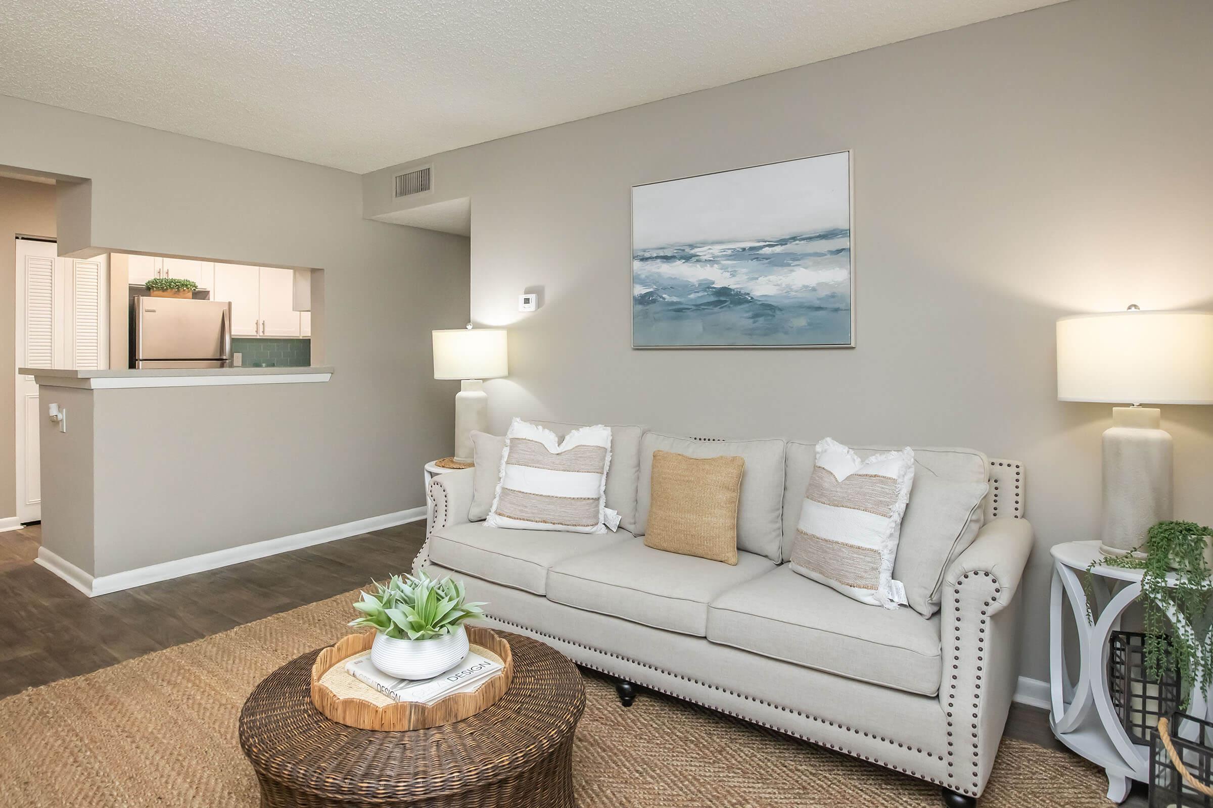 Cozy living room featuring a light gray sofa with decorative pillows, a round woven coffee table with a plant centerpiece, and two table lamps. The walls are a soft neutral color, and a large abstract painting is hung above the sofa. In the background, an open kitchen area is partially visible.