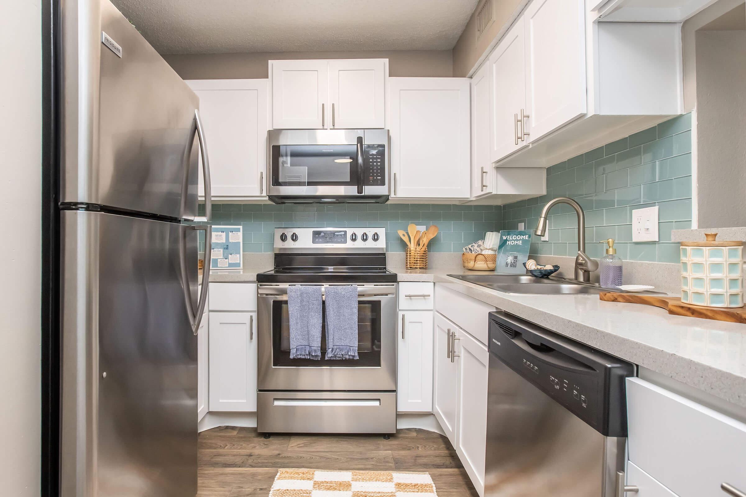 A modern kitchen featuring stainless steel appliances, including a refrigerator, oven, microwave, and dishwasher. The cabinets are white, and the backsplash is light blue tiles. A countertop with a cutting board and utensils is visible, along with a small area rug on the floor.