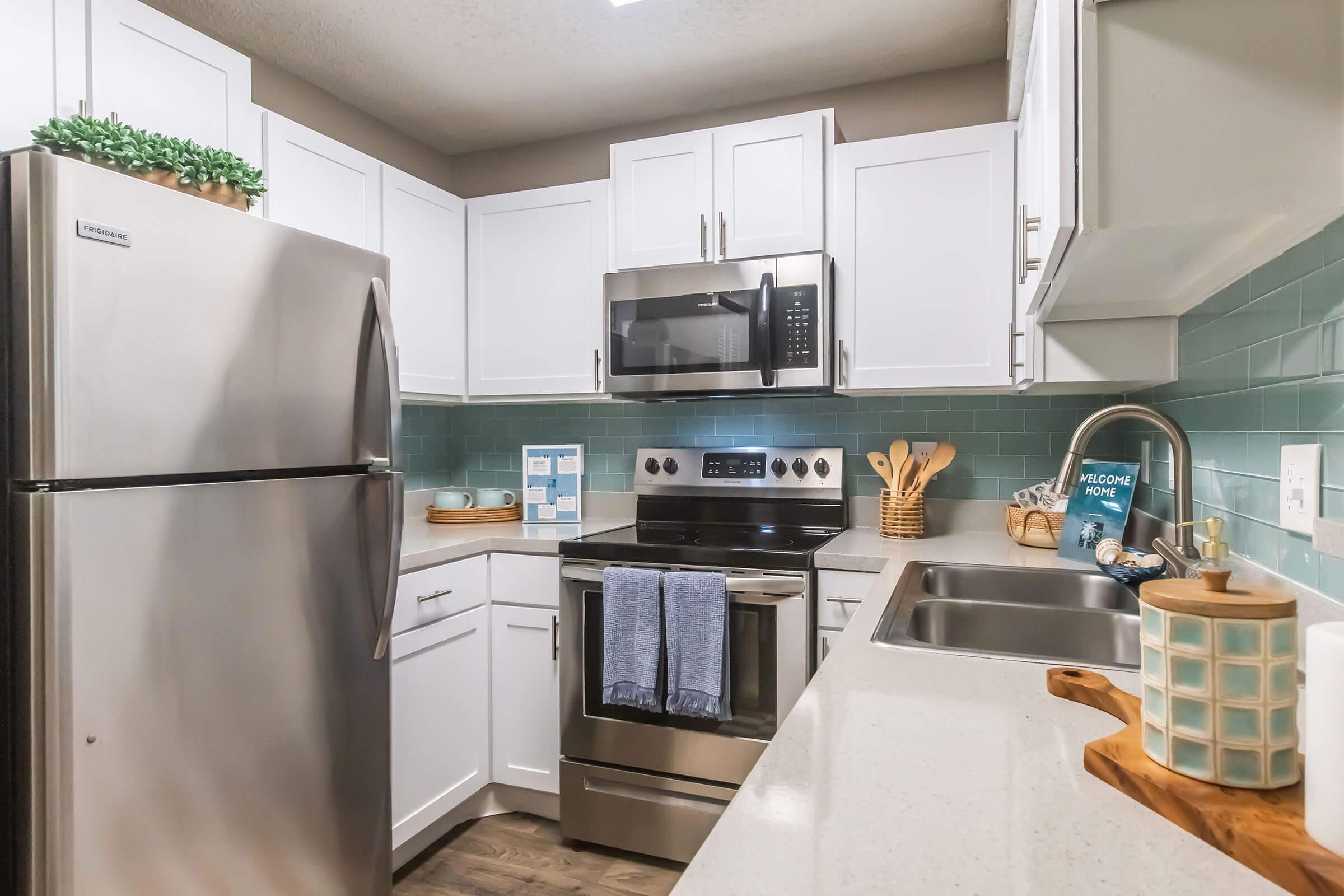Modern kitchen featuring white cabinetry, stainless steel appliances, and a teal tile backsplash. The space includes a refrigerator, microwave, oven, and sink with wooden utensils and decorative items on the counter, creating a bright and welcoming atmosphere.