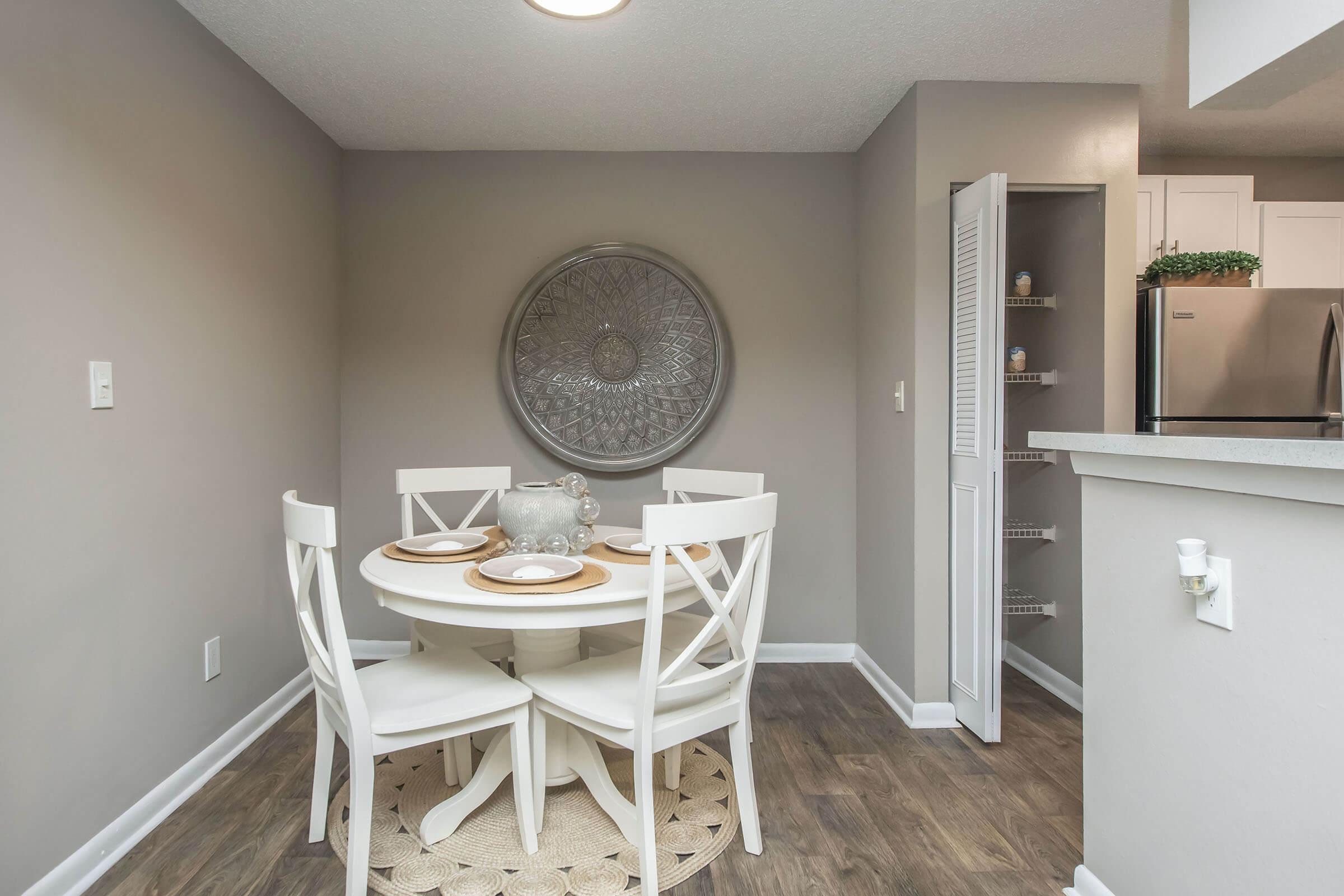 A cozy dining area featuring a round white table with four chairs, set with placemats and tableware. A decorative round wall art piece hangs behind the table, and a kitchenette is visible in the background, showcasing stainless steel appliances and shelving. The walls are painted in neutral tones, creating a warm atmosphere.