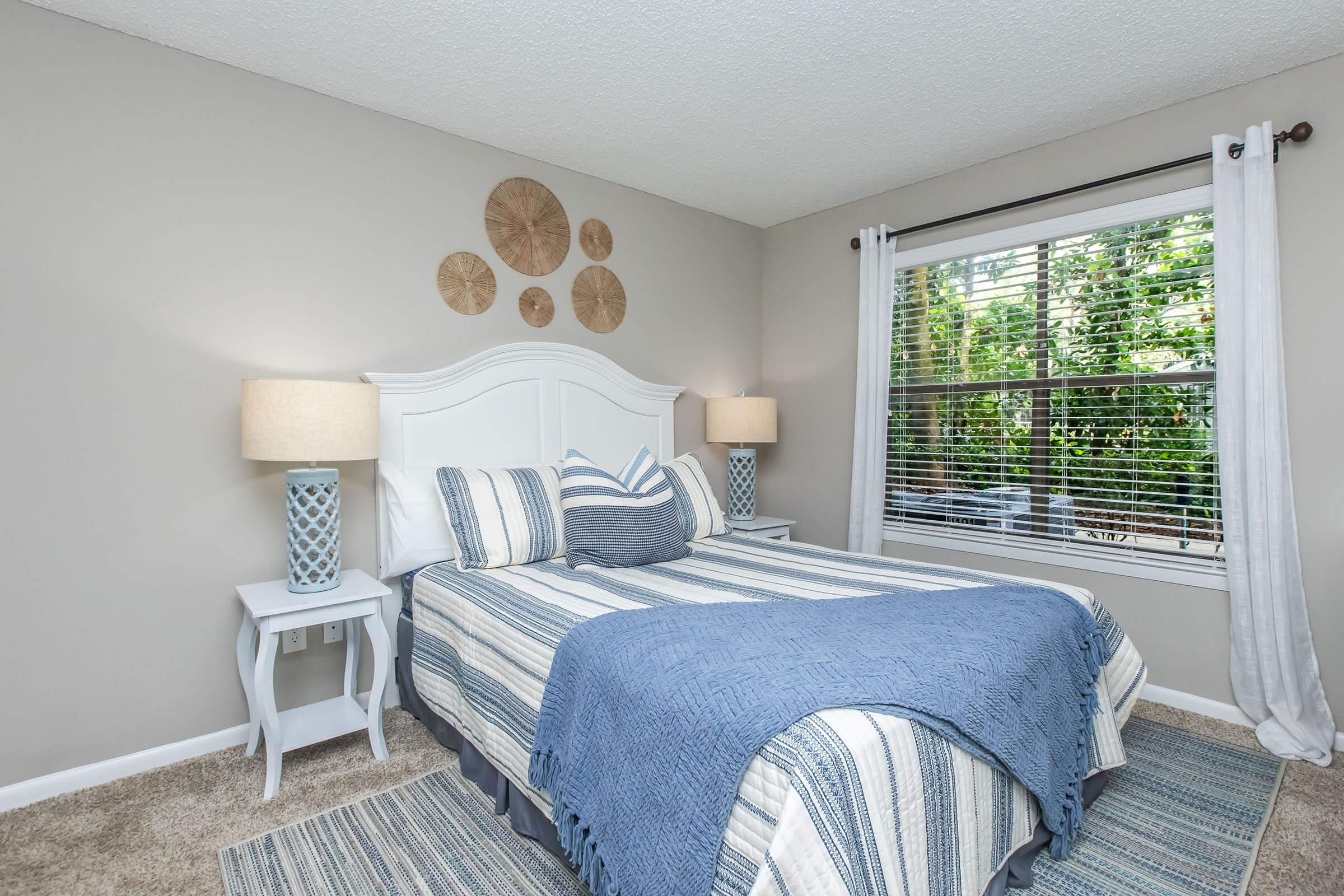 A cozy bedroom featuring a queen-sized bed with striped blue and white bedding. There are two bedside tables with decorative lamps, and above the bed, circular wall decor in a natural finish. The room has a window with sheer curtains, allowing natural light to filter in, and the walls are painted a soft gray.