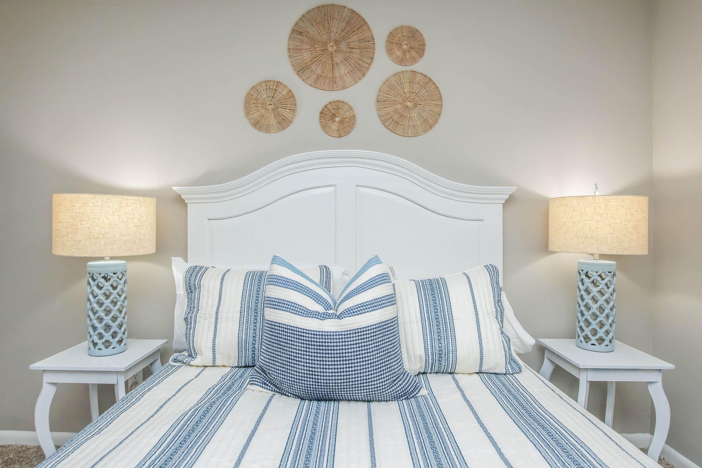 A neatly made bed with a white headboard and blue and white striped bedding. Two decorative lamps with light shades are positioned on either side of the bed. Above the bed, three round wall decorations made of natural materials add a touch of decor to the room's neutral walls.