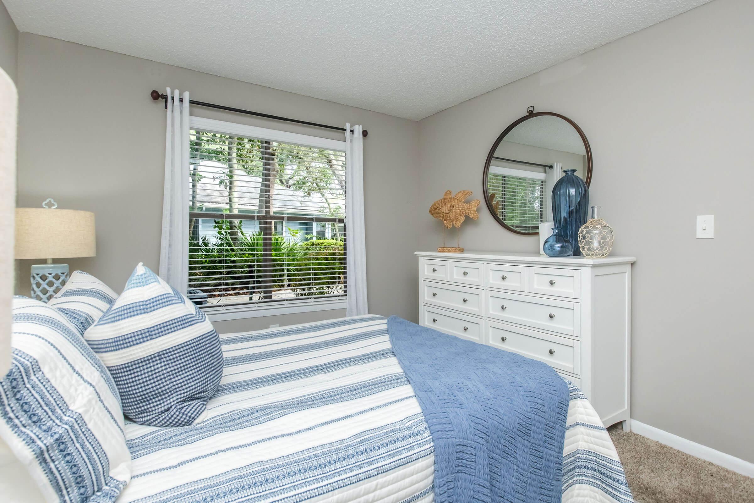 A cozy bedroom featuring a white dresser, a striped blue and white bedspread, and decorative pillows. Natural light filters through a window adorned with sheer curtains, revealing a glimpse of greenery outside. Decorative items, including a large fish sculpture and glass accents, enhance the room's aesthetic.