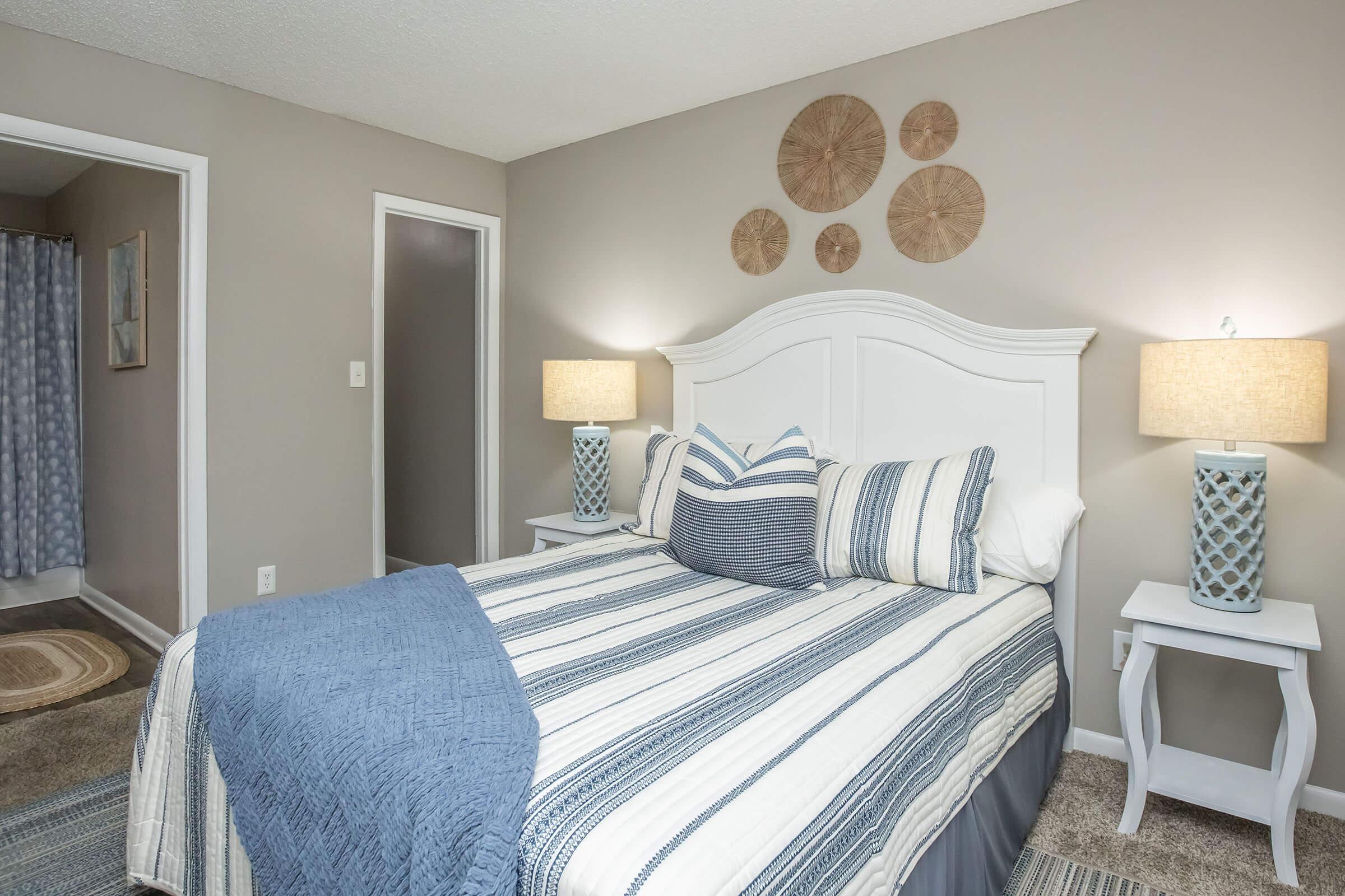 A cozy bedroom featuring a white bed with blue and striped bedding, complemented by decorative pillows. There are two bedside tables with lamps, and three circular wall decorations above the headboard. A doorway visible on the left leads to a bathroom with a shower curtain, and the room has soft carpet flooring.
