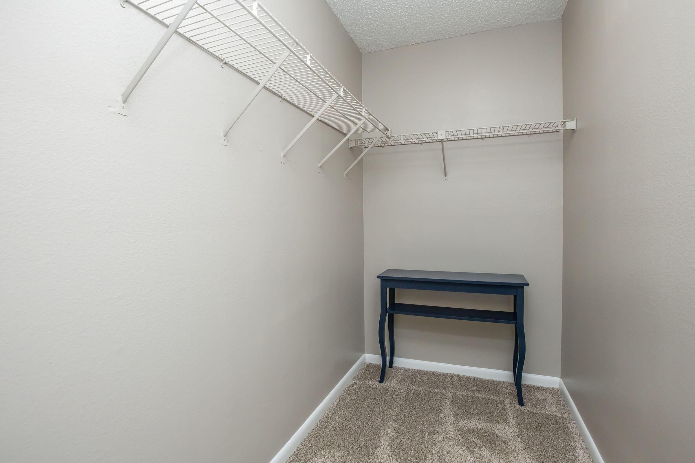 Empty closet space with light gray walls and a beige carpet. A wire shelf is attached to the wall on the left, and a small blue table is positioned in the center. The space is well-lit and organized, suitable for storage or as a small workspace.