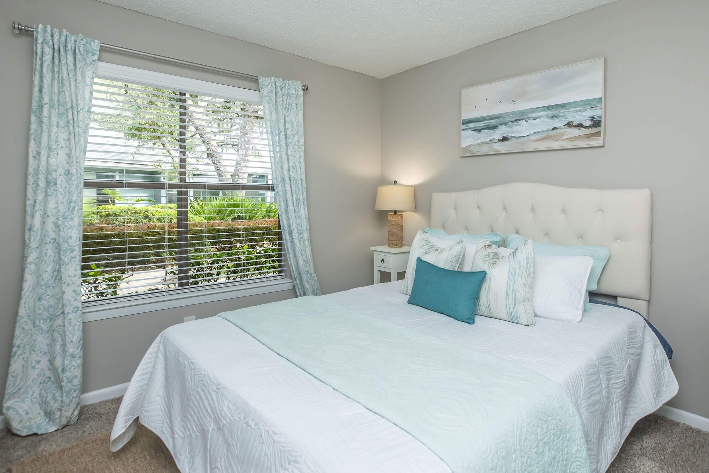 A cozy bedroom featuring a queen-sized bed with light blue and white bedding. There are two decorative pillows on the bed. Natural light pours in from a window with sheer curtains, showcasing a green outdoor view. A bedside table with a lamp and a piece of ocean-themed artwork adorn the gray walls.