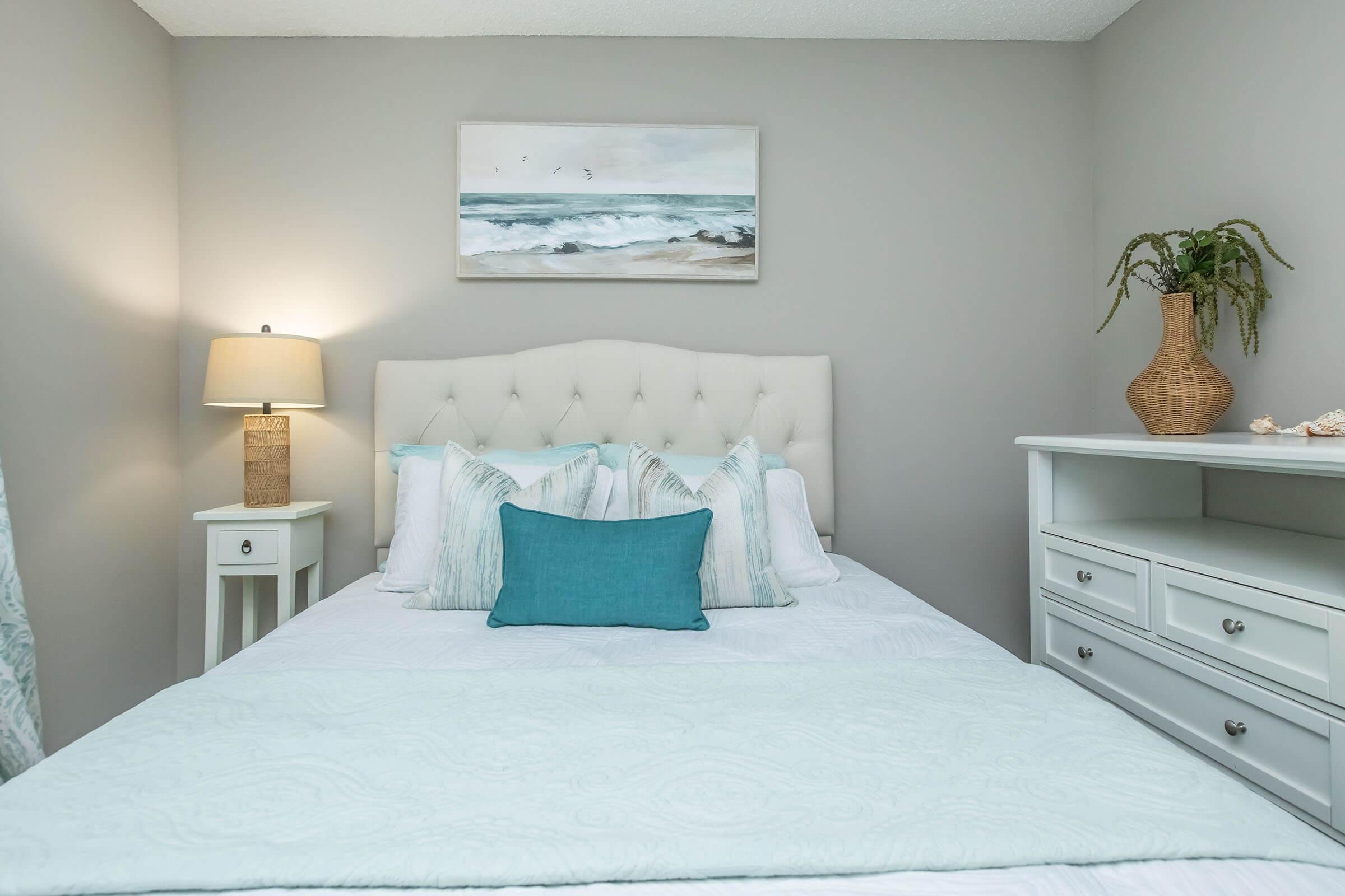 A cozy bedroom featuring a light-colored bed with a decorative aqua-blue pillow and a soft quilt. There's a framed ocean painting on the wall, a bedside lamp on a white nightstand, and a white dresser with drawers. A potted plant adds a touch of greenery to the calming, neutral decor.