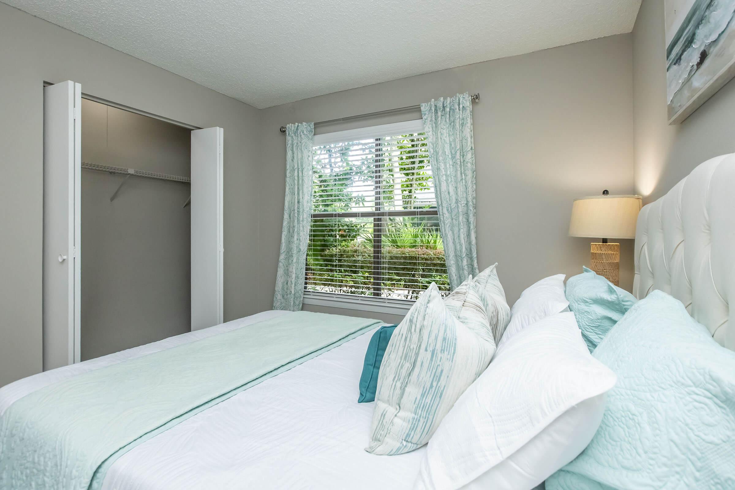 A cozy bedroom featuring a white bedspread with light blue decorative pillows. Natural light pours in through a window with sheer curtains, revealing greenery outside. There's a bedside lamp on a nightstand and a closet with sliding doors on the left. The walls are painted a soft gray, adding to the tranquil atmosphere.