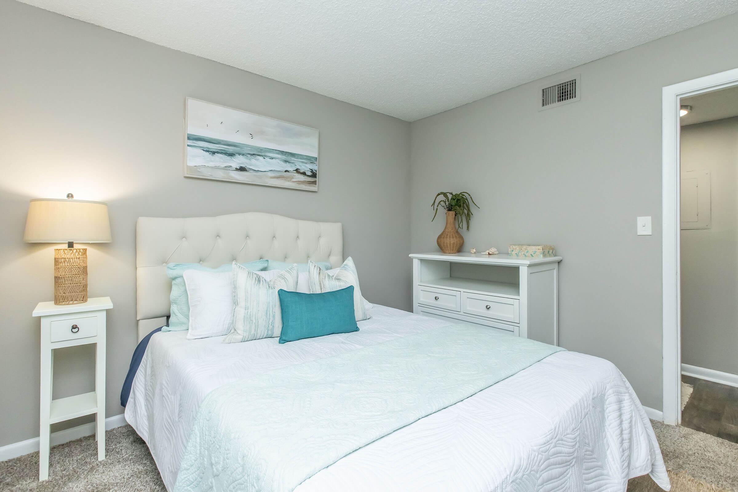 A cozy bedroom featuring a light-colored tufted headboard with a pale blue and white bedding set. A bedside table with a lamp sits next to the bed, and a white dresser is against the wall. A decorative plant and a piece of ocean-themed artwork adorn the space, which has soft gray walls and a welcoming ambiance.